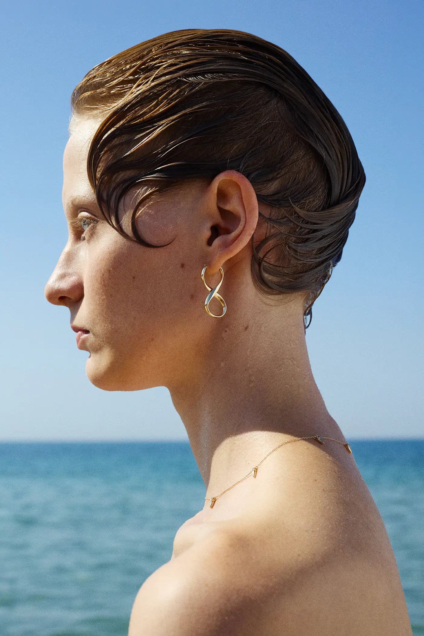 A woman with slicked-back wet hair stands against clear blue sky and a calm sea view, wearing a pair of sculptural gold earrings and a fine gold necklace with small diamond charms.
