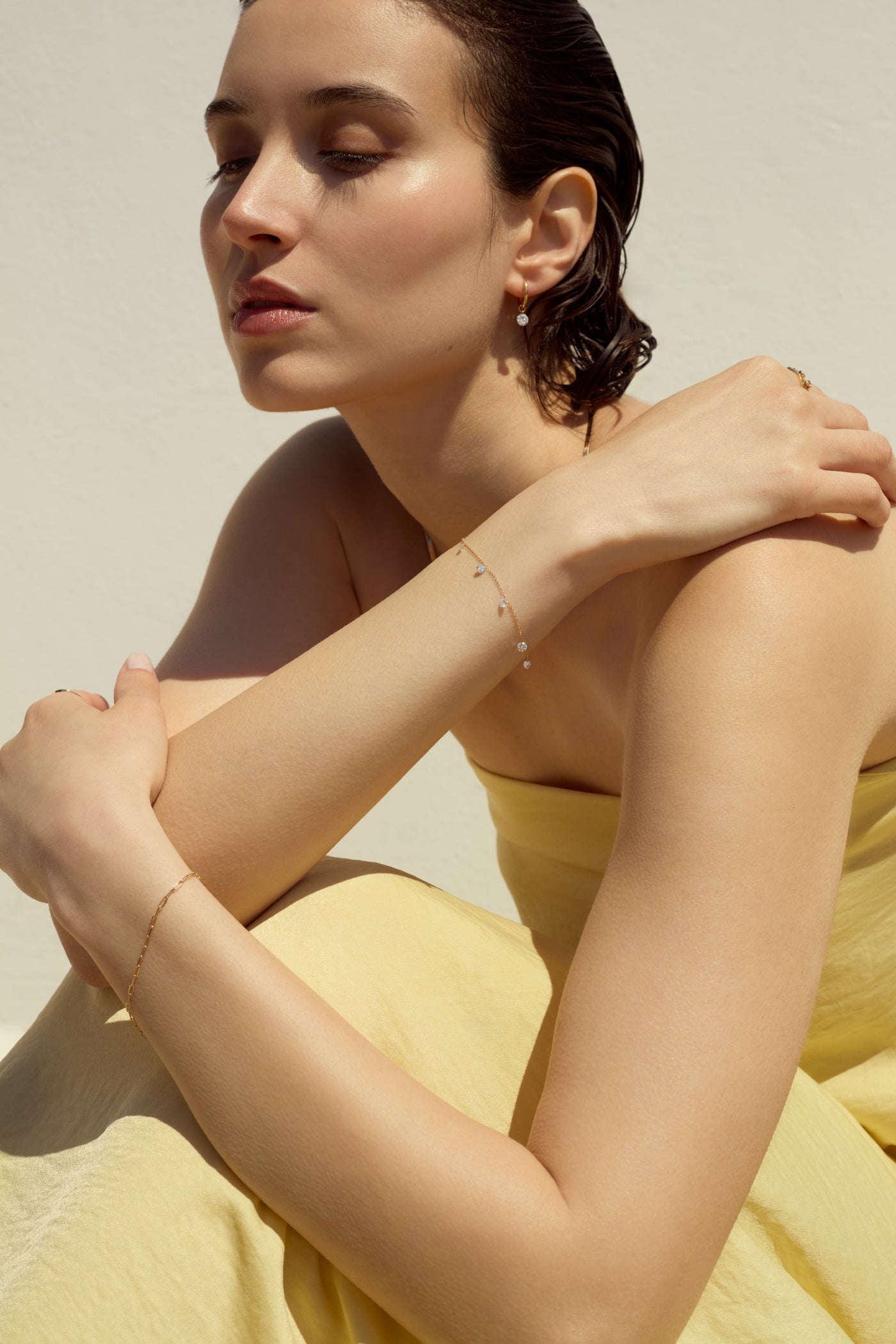 Portrait of a woman in a light yellow dress, wearing a delicate 14k yellow gold bracelet with five dangling diamonds on one hand, a 14k gold paperclip style bracelet on her other hand, and a small gold hoop that features one drop diamond. 