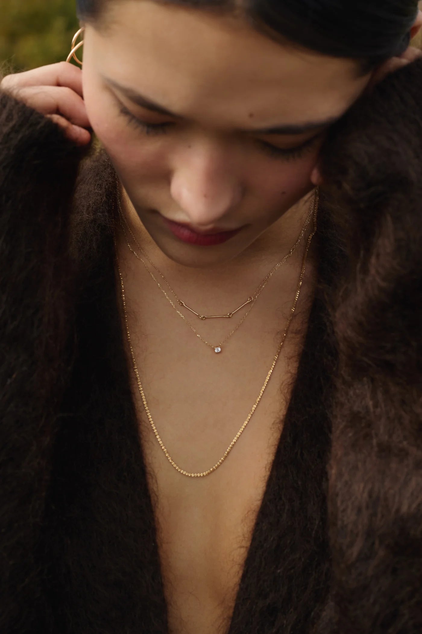 Portrait of a woman wearing a long 14k gold chain necklace and a 14k gold necklace with a dainty diamond, stacked with a shorter 14k gold necklace with links in a natural backround, styled with a brown fur coat. — Enea