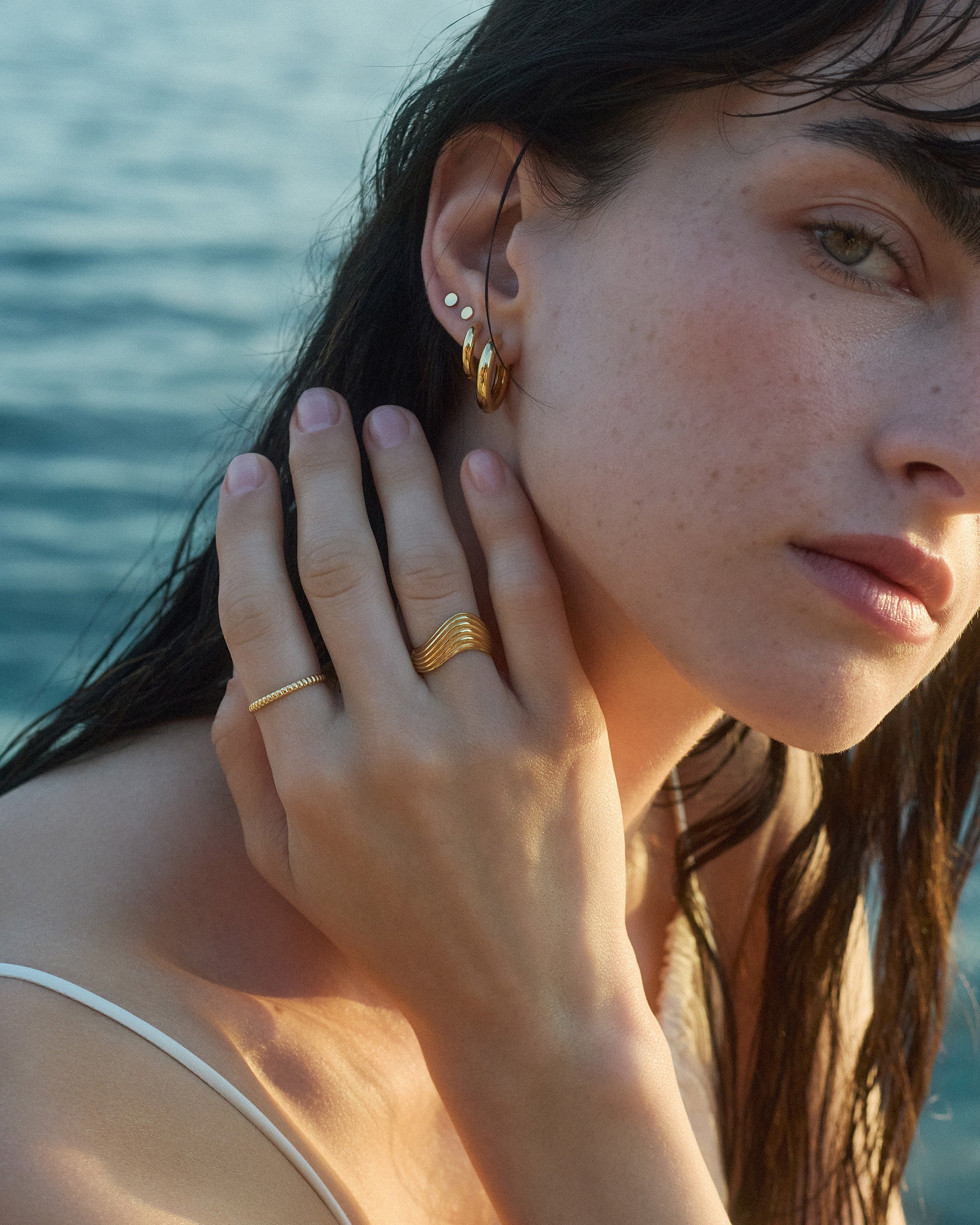 Woman wearing gold earrings, a fine textured band and the Kion III Ribbed Wave Ring by ENEA with a blurred water background
