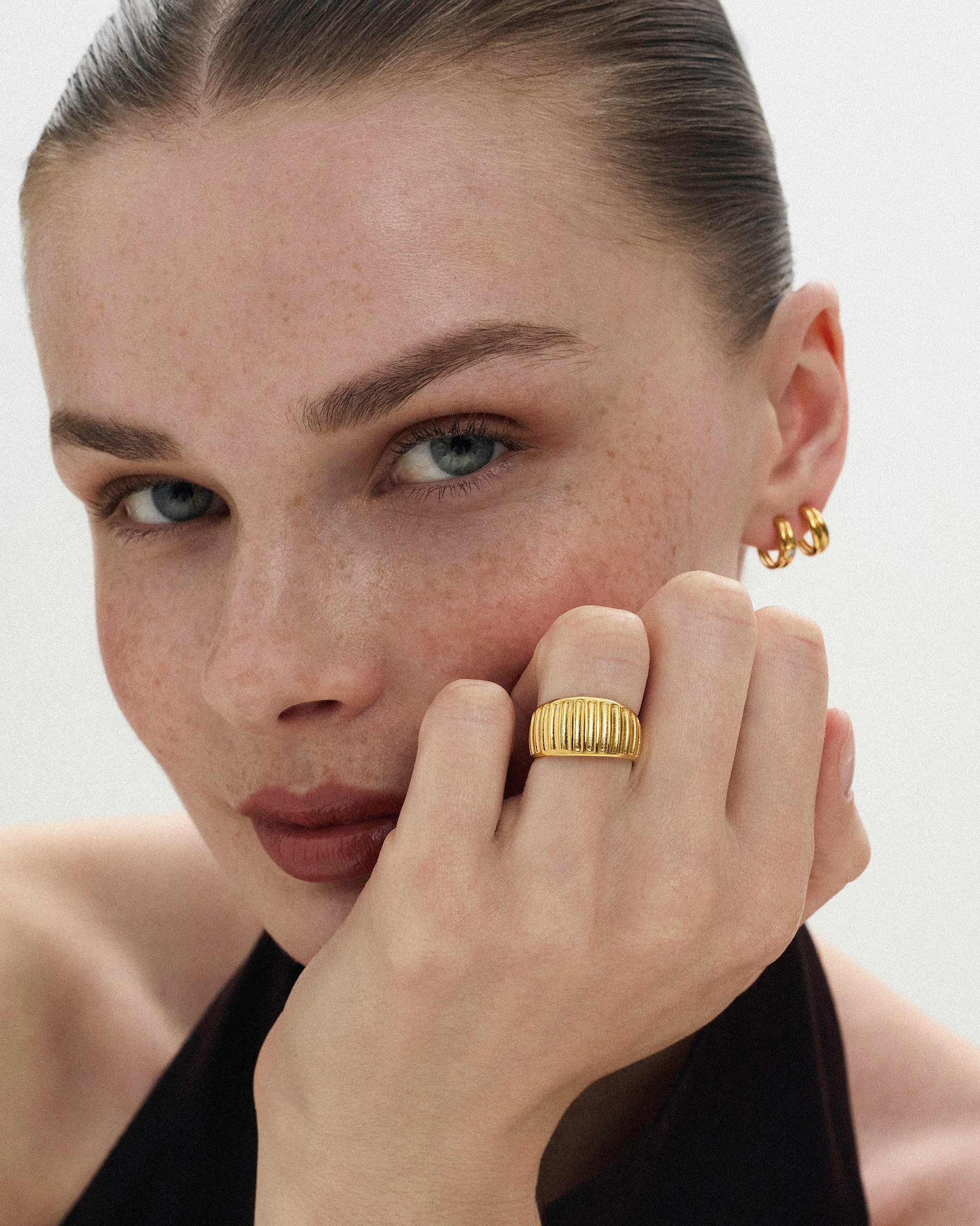 Woman resting her hand on her face wearing solid gold jewelry including small hoop earrings and a ribbed ring