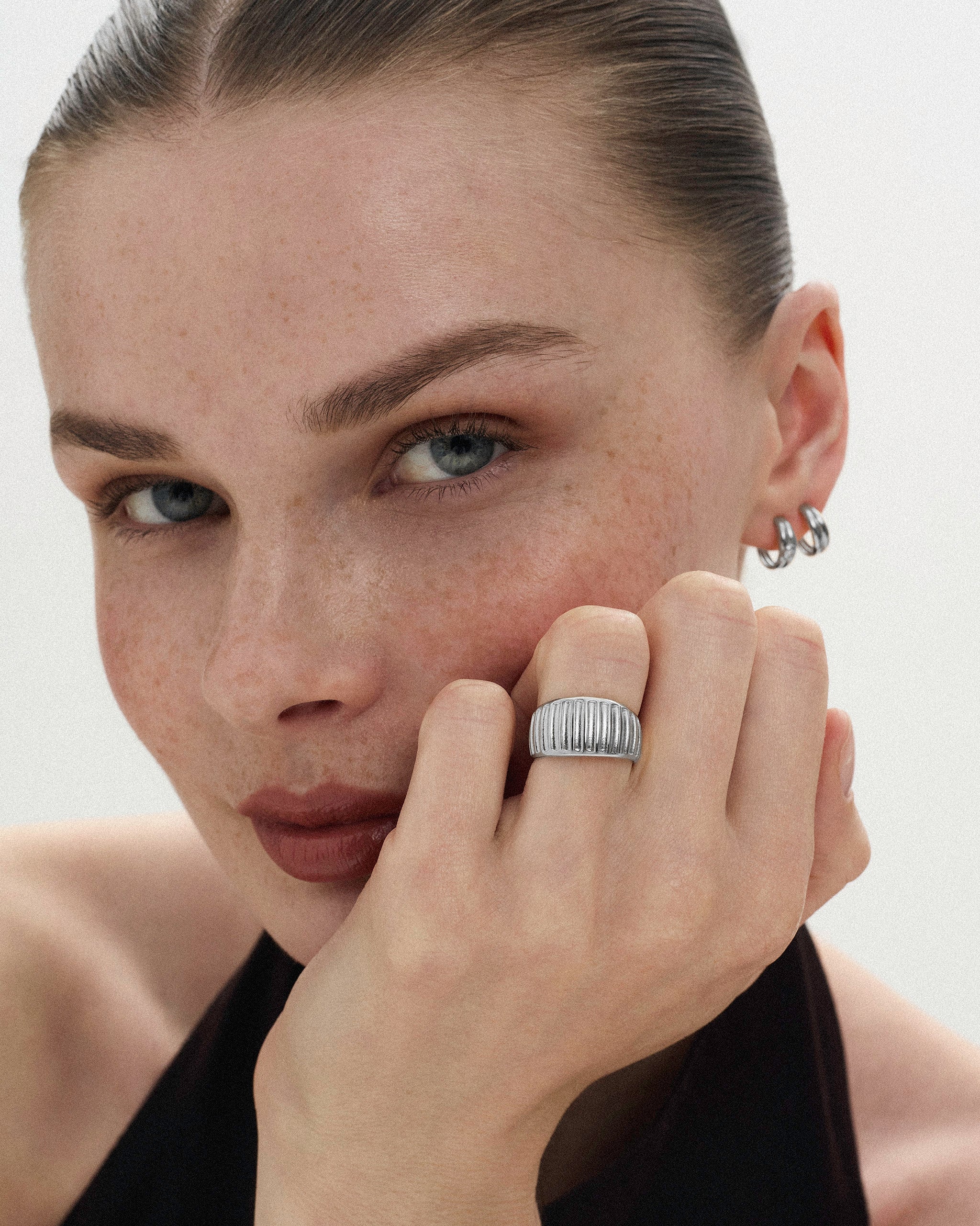 Woman resting her hand on her face wearing silver jewelry including small hoop earrings and a ribbed ring