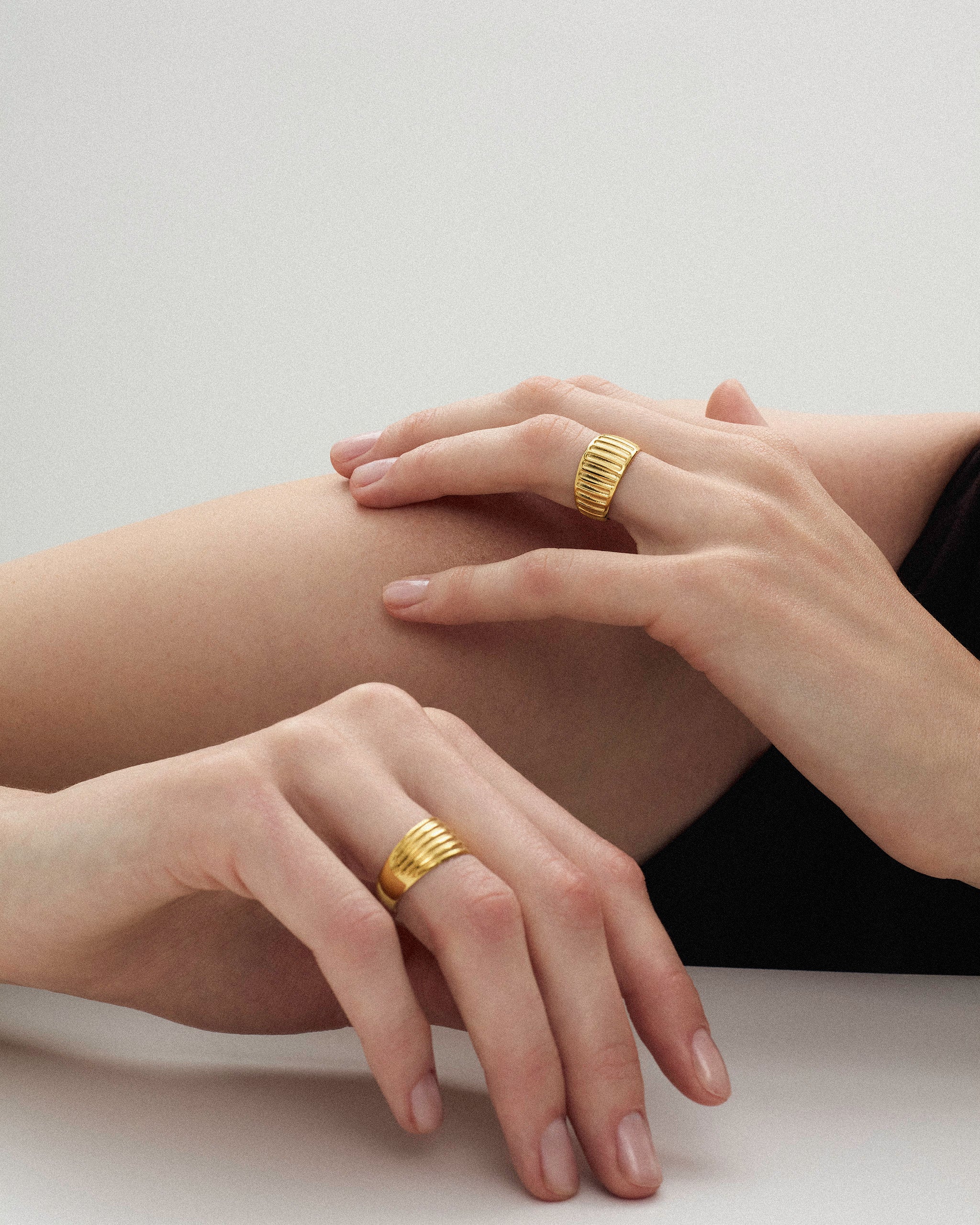 Close-up of a woman's hands styled with ribbed solid gold rings on a neutral background