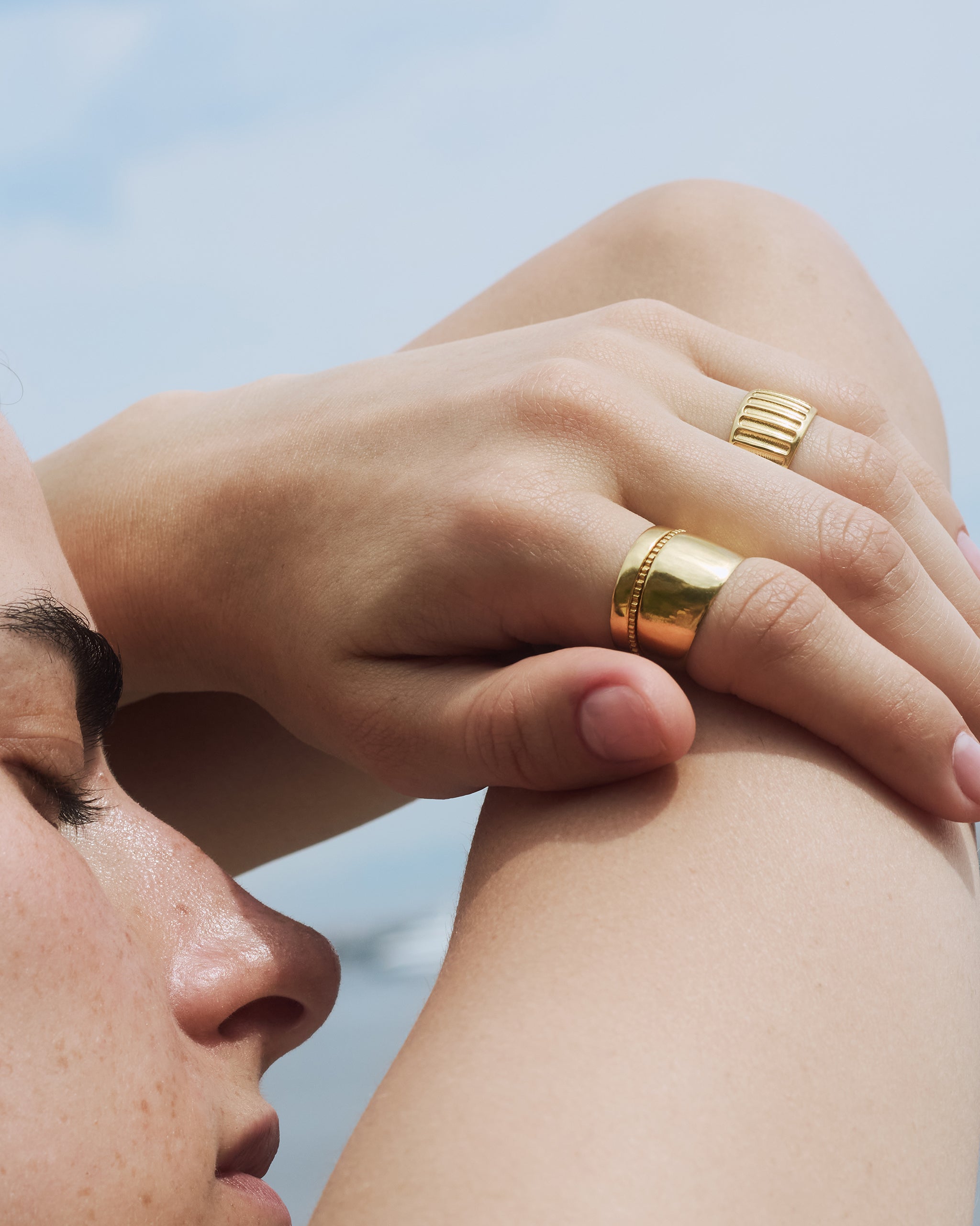 A woman resting her hand on her arm with thick solid gold bands styled on one hand against a light blue background