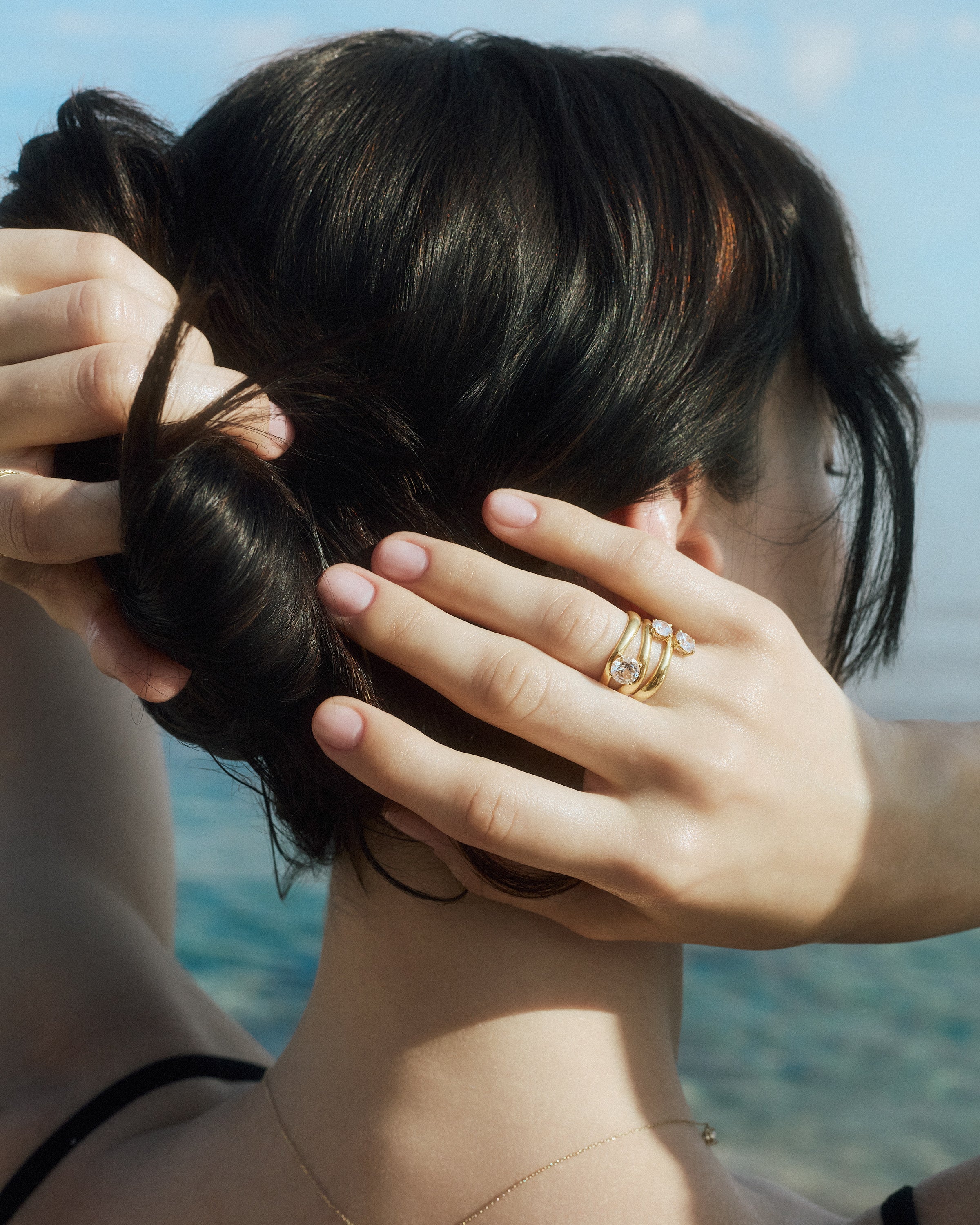 back view of a woman touching her hair with her hand wearing the kyma solitaire rings in solid yellow gold in 0.50ct, 0.75ct and 1ct sizes on a sea background