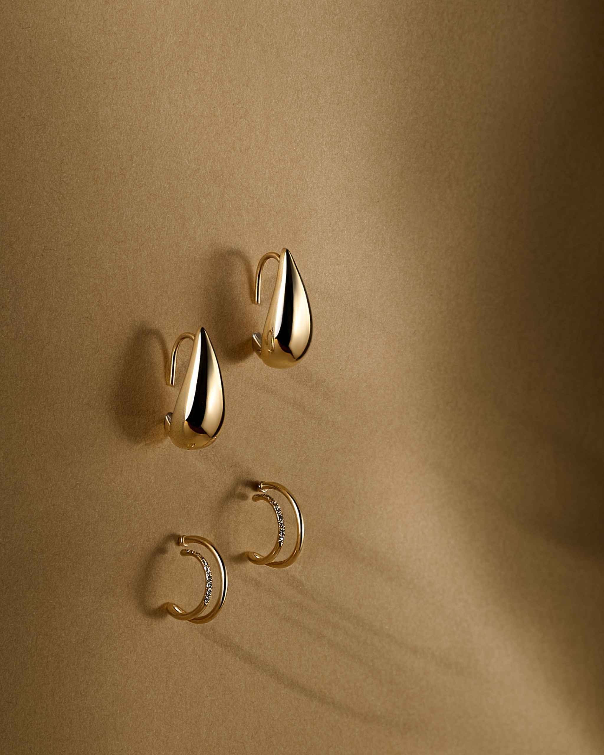 Two pairs of solid gold hoop earrings displayed on a warm beige background: one pair of teardrop-shaped hoops and a second pair of slim curved hoops with small diamonds, arranged diagonally with soft shadows.