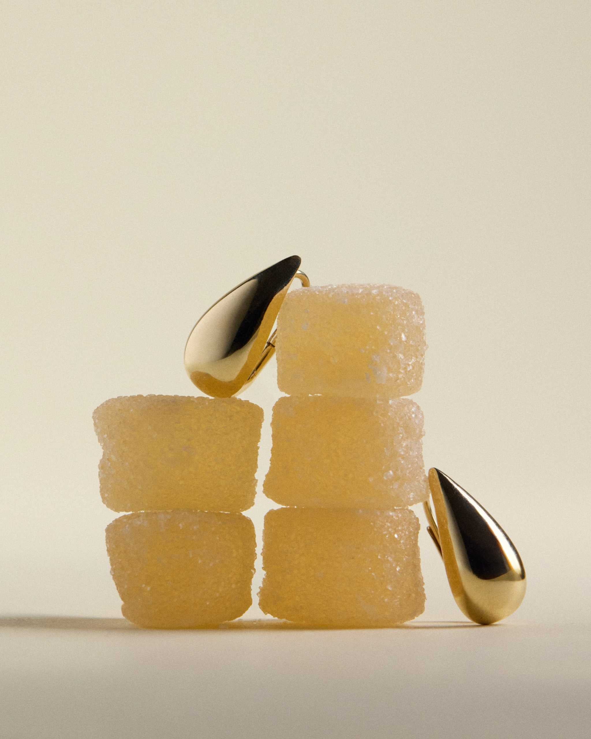 A pair of solid gold teardrop-shaped earrings displayed on a stack of yellow sugar cubes against a warm cream background.