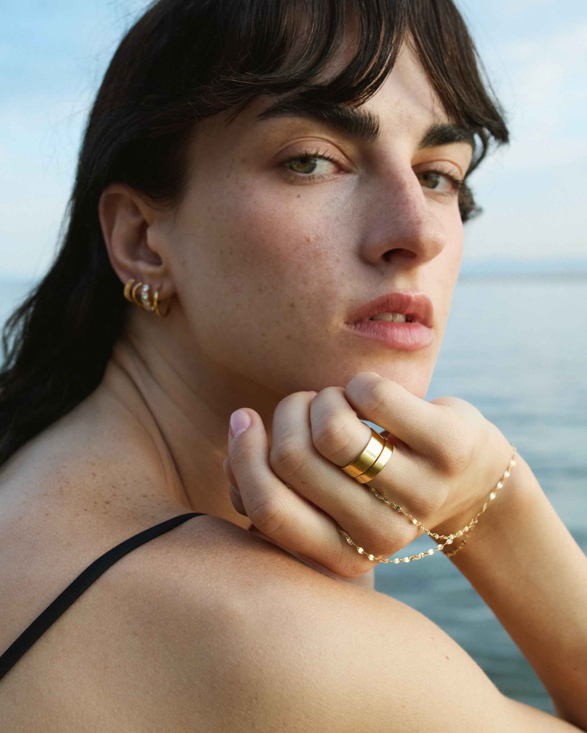 The Lien Hand Chain Marine Bracelet drape from a woman's hand styled with thick gold bands and hoop earrings on blurred sea background