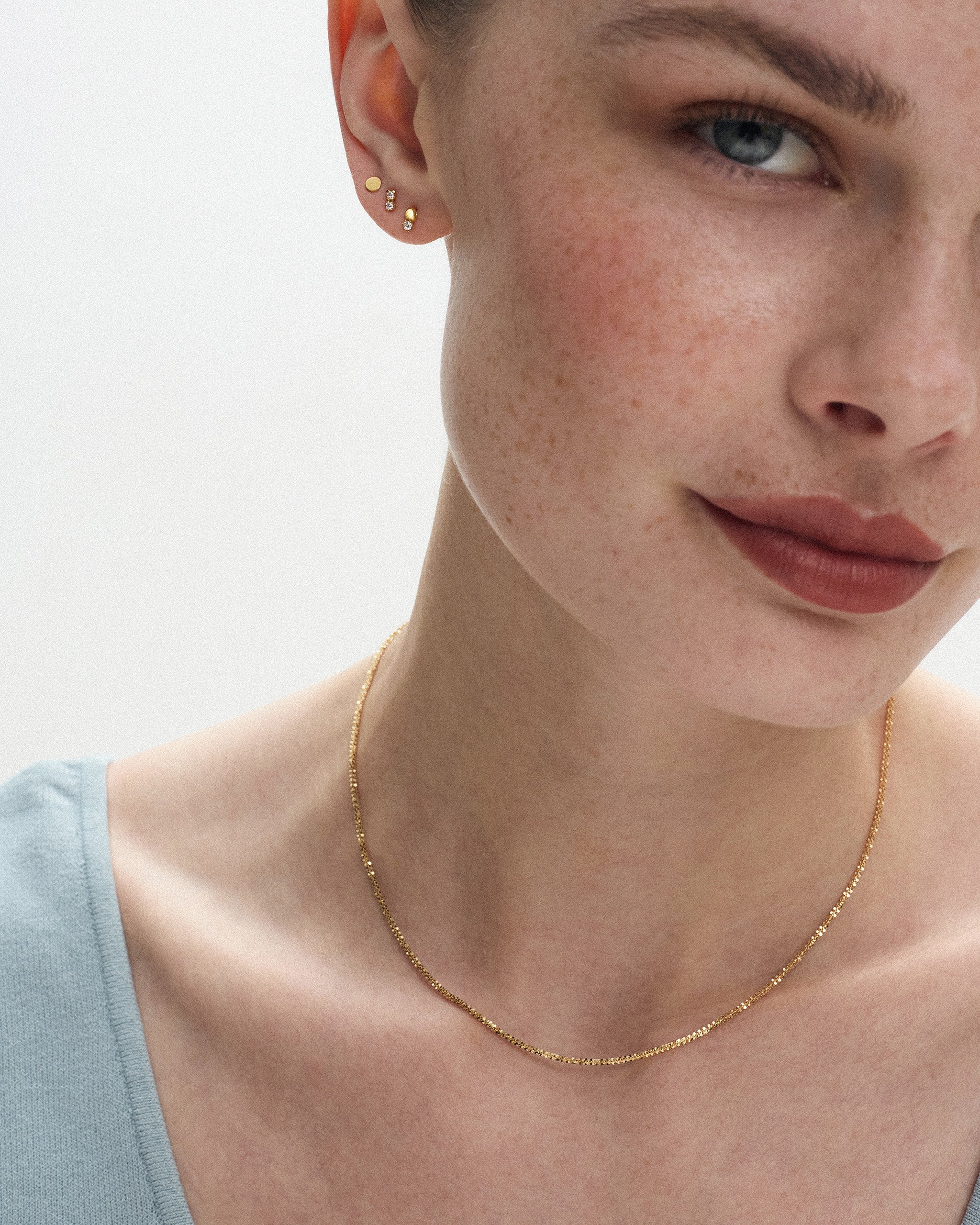 Woman wearing the Lumière Sparkle Chain Necklace in 14k yellow gold at the collarbone with three small gold studs in her ear. ENEA Studio