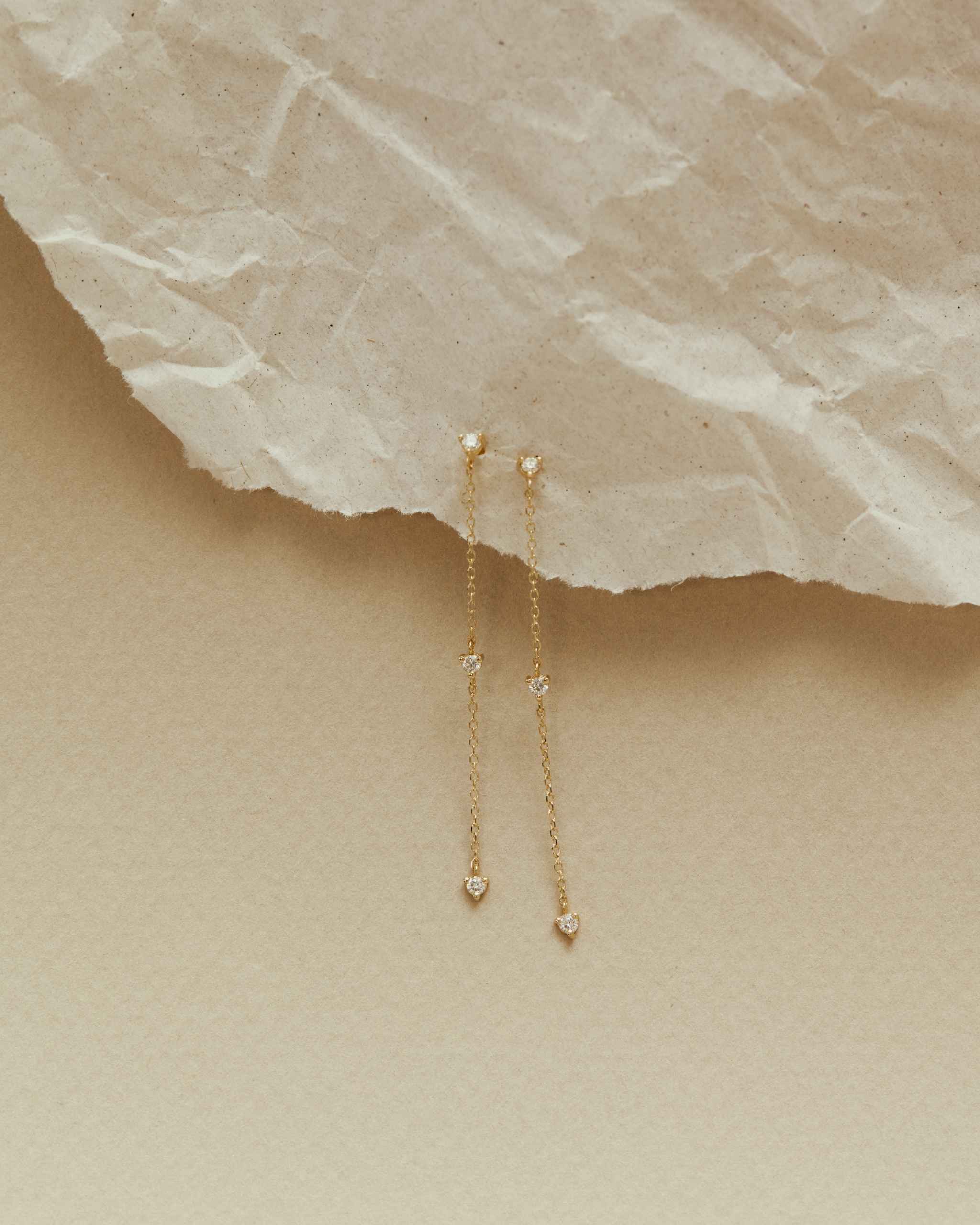 a pair of 14k Gold Nema Trio Diamond Drop Earrings with three diamonds laid on crumpled paper on a soft beige surface