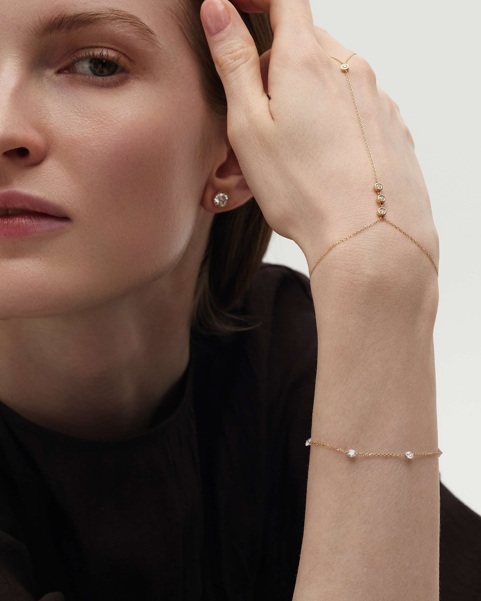 woman resting her hand on her face wearing a diamond stud earring, a dainty gold hand chain with our bezel diamonds and a delicate nude diamond station bracelet