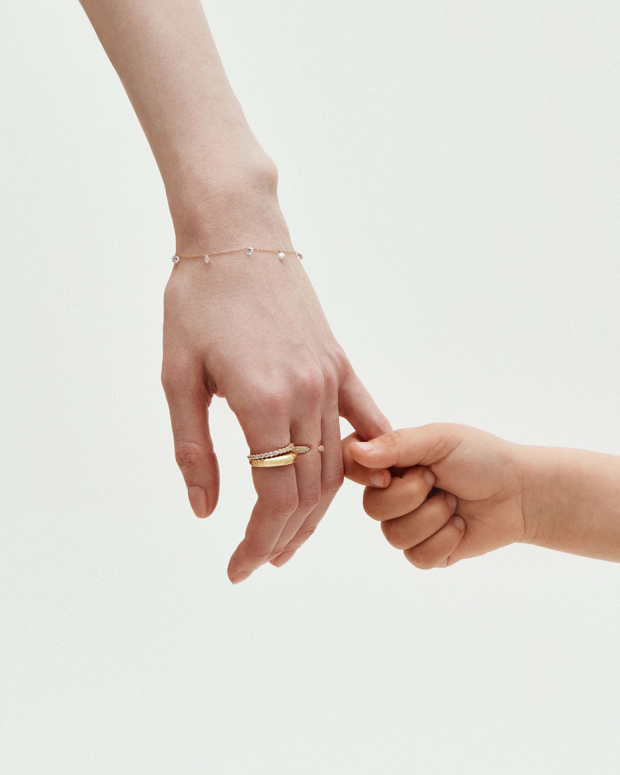 A women's hand holds a toddler's hand. The woman's hand is styled with solid gold, diamond-accented bands and a delicate gold bracelet with drop diamonds.
