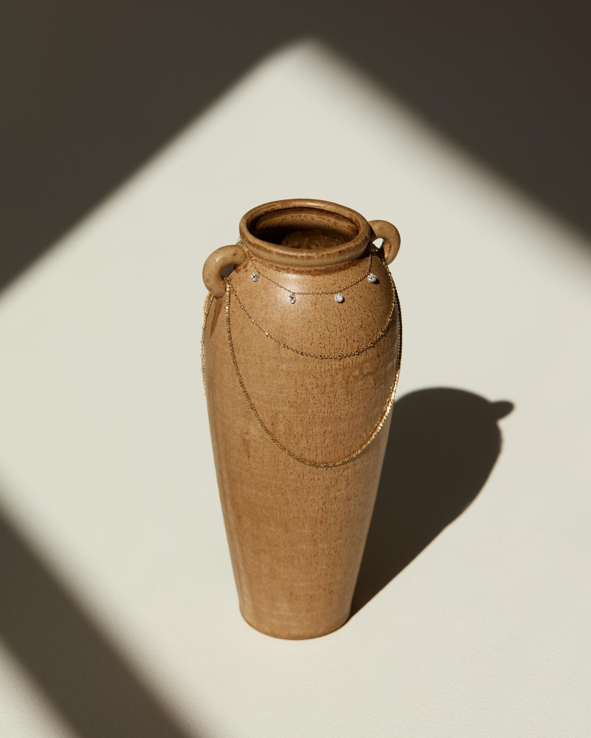A tall, slender ceramic vase with a warm brown earthy glaze, featuring two side handles and a decorative chain detail, set against a minimalist background with dramatic diagonal shadows.