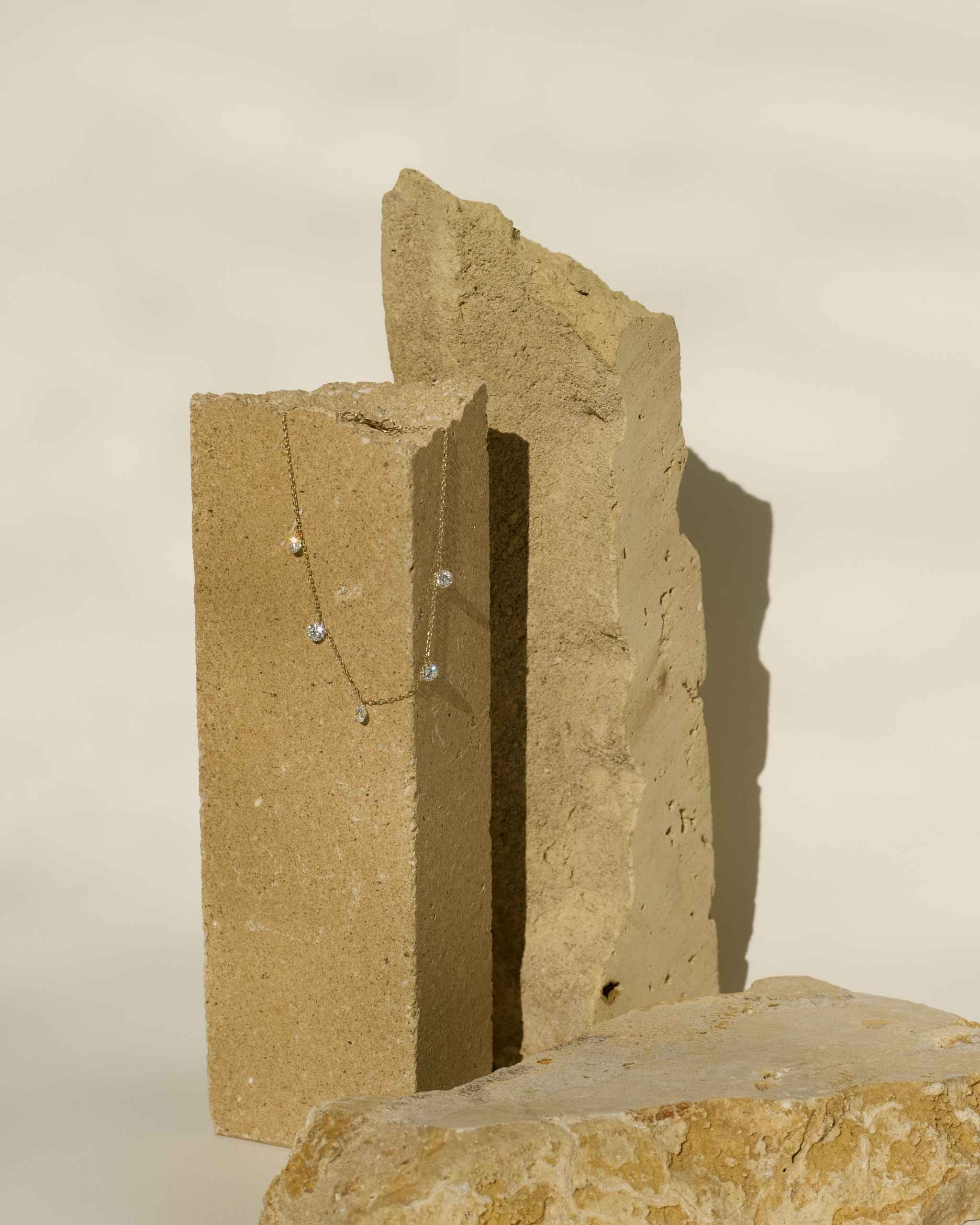A delicate solid gold bracelet with dangling diamonds draped over two textured stone blocks against a warm off-white background.