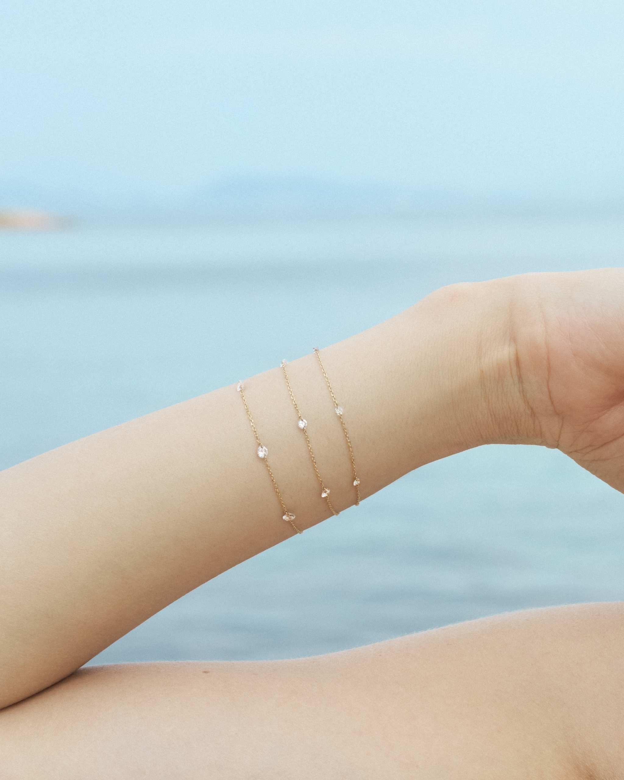 Close-up of a woman wrist wearing three gold diamond chain bracelets with delicate nude diamonds of different sizes on a calm sea background