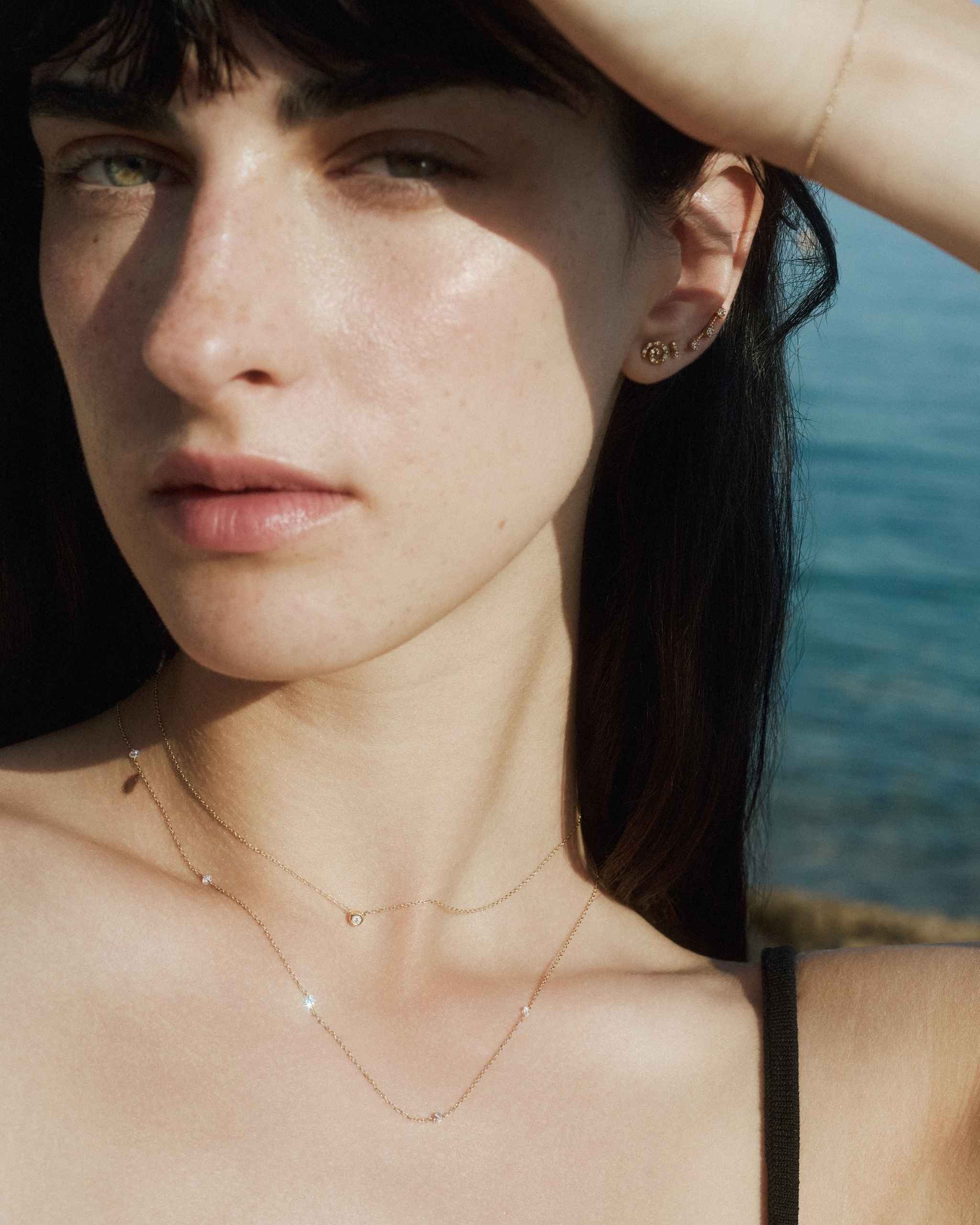 a woman under soft sunlight wearing a solid gold diamond station necklace with four diamonds layered with a petite bezel diamond necklace and dainty diamond studs on a calm sea background