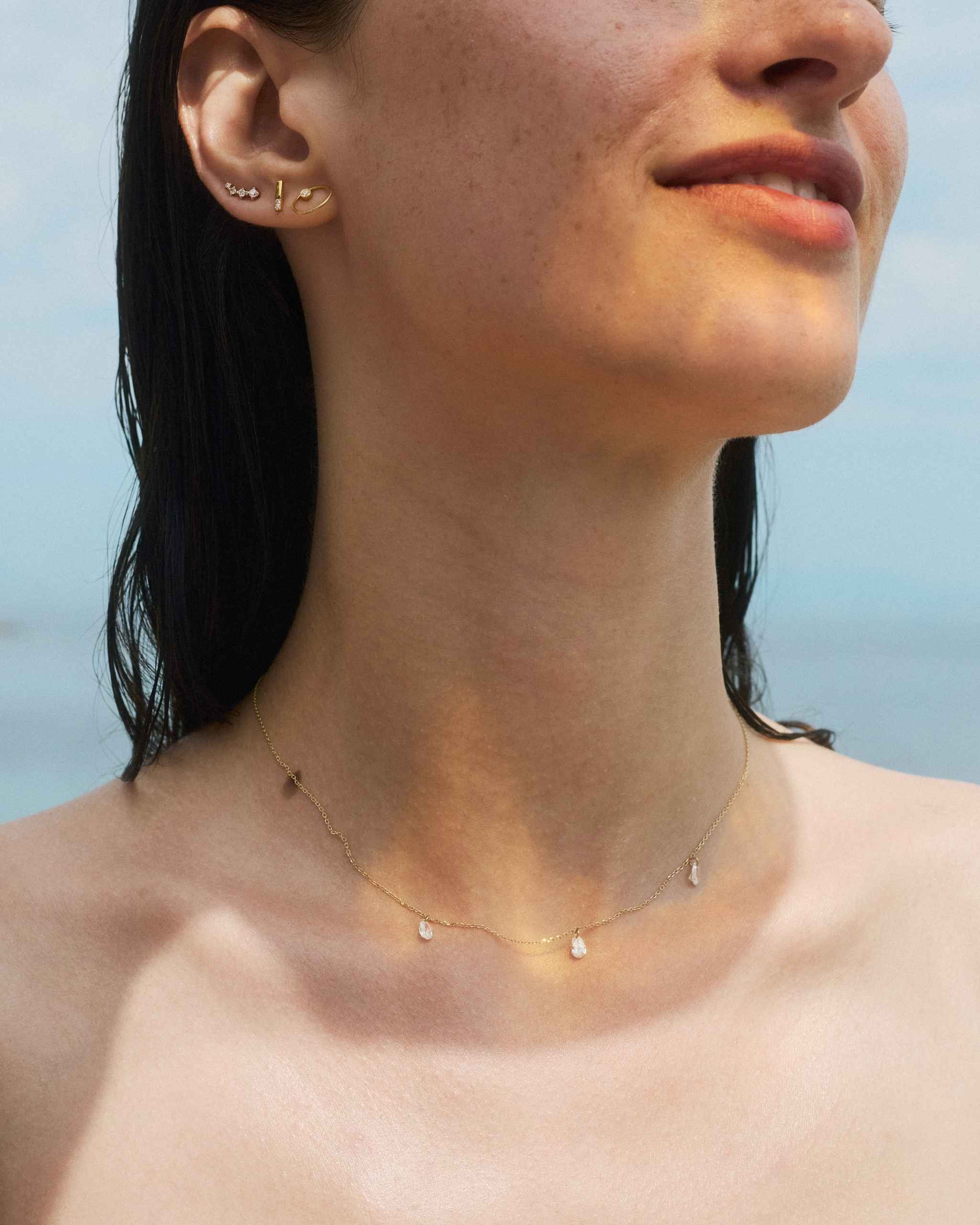 woman wearing the solid gold pear diamond necklace styled with delicate gold and diamond studs on a blurred sea background