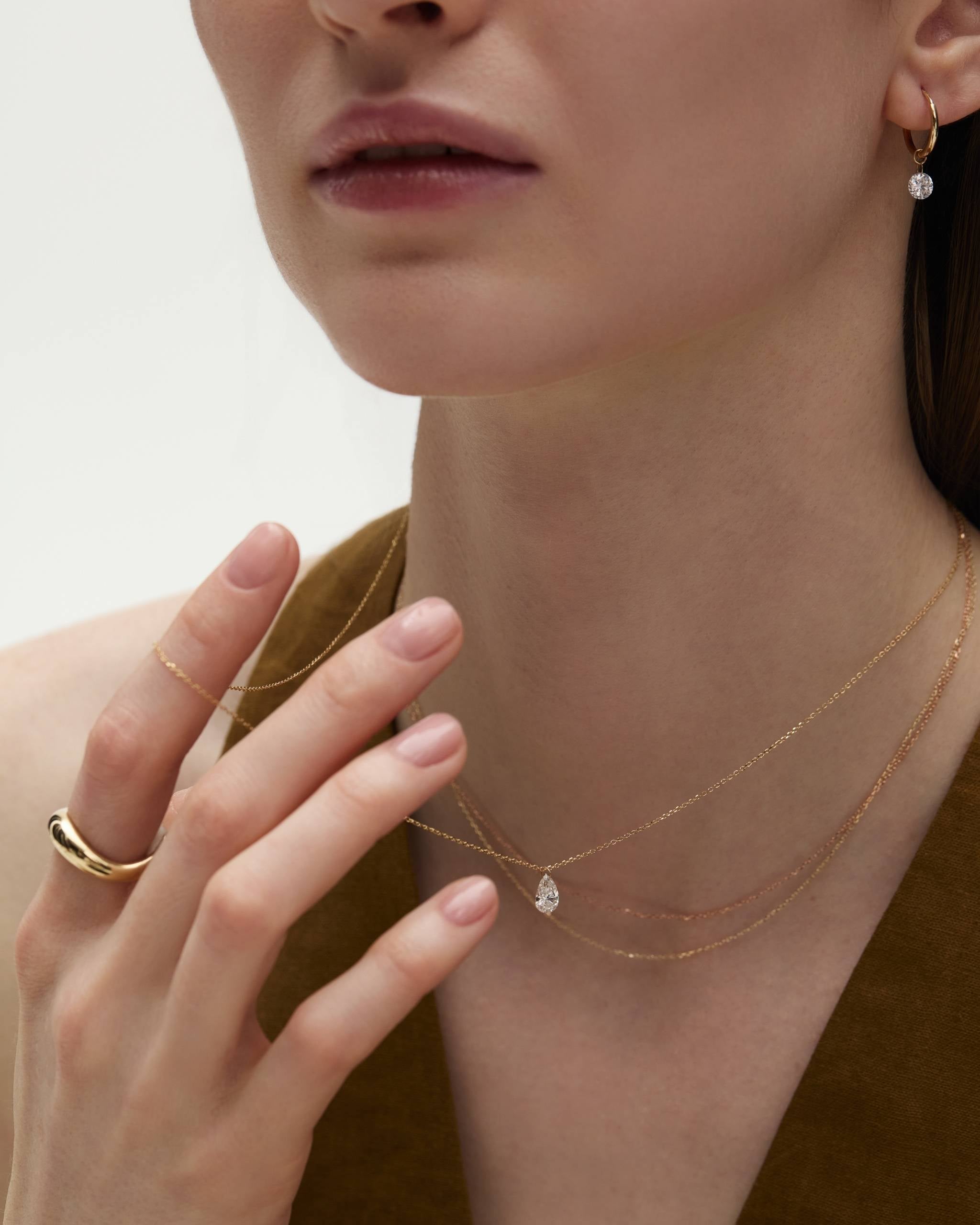 model wears a 14k gold necklace with a pear-shaped solitaire nude diamond styled with a thick curved band and a gold hoop with a dangling nude diamond