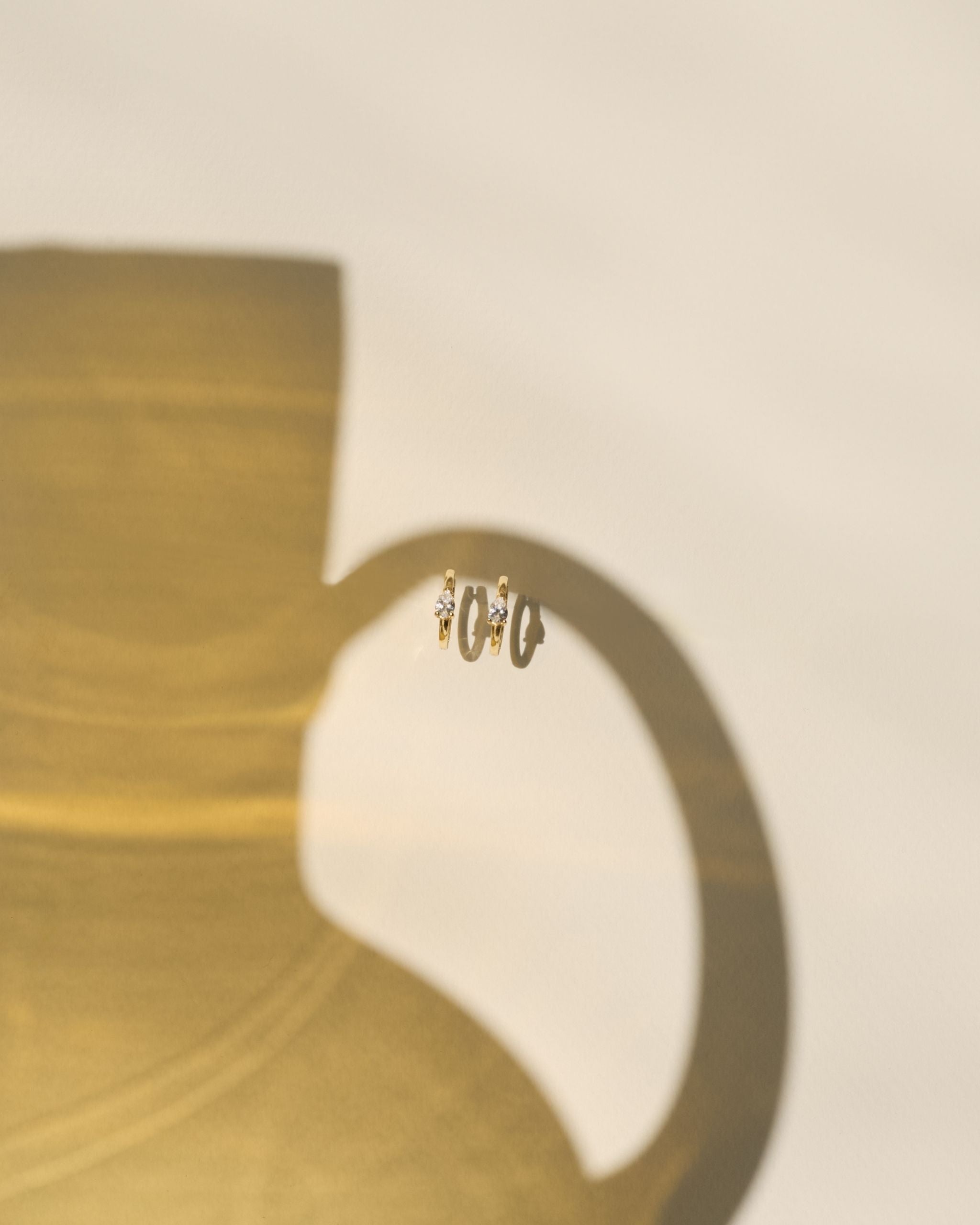A pair of small gold hoop earrings with pear shaped stones, shown against a warm beige background with a large soft shadow cast by a gold-toned object.