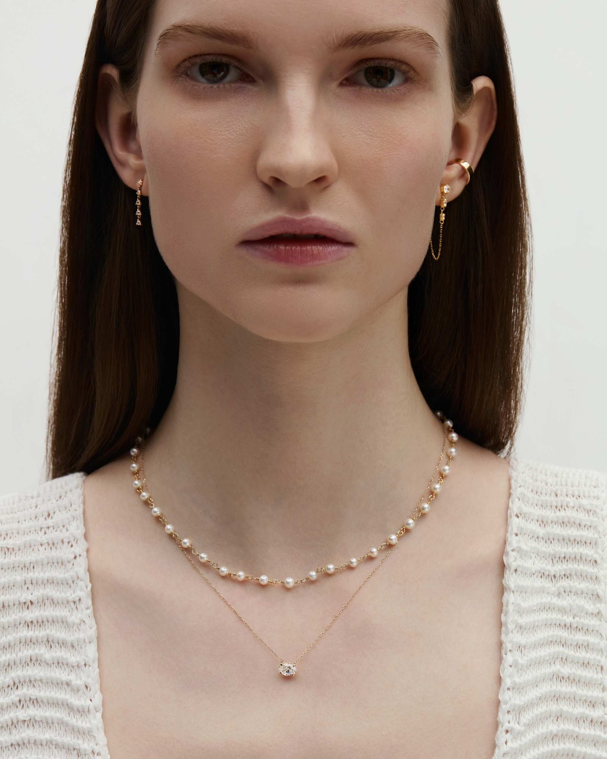 front view of a woman wearing a 14k gold pearl wrap necklace layered with a longer diamond solitaire necklace and styled with dainty chain and diamond earrings on a white background