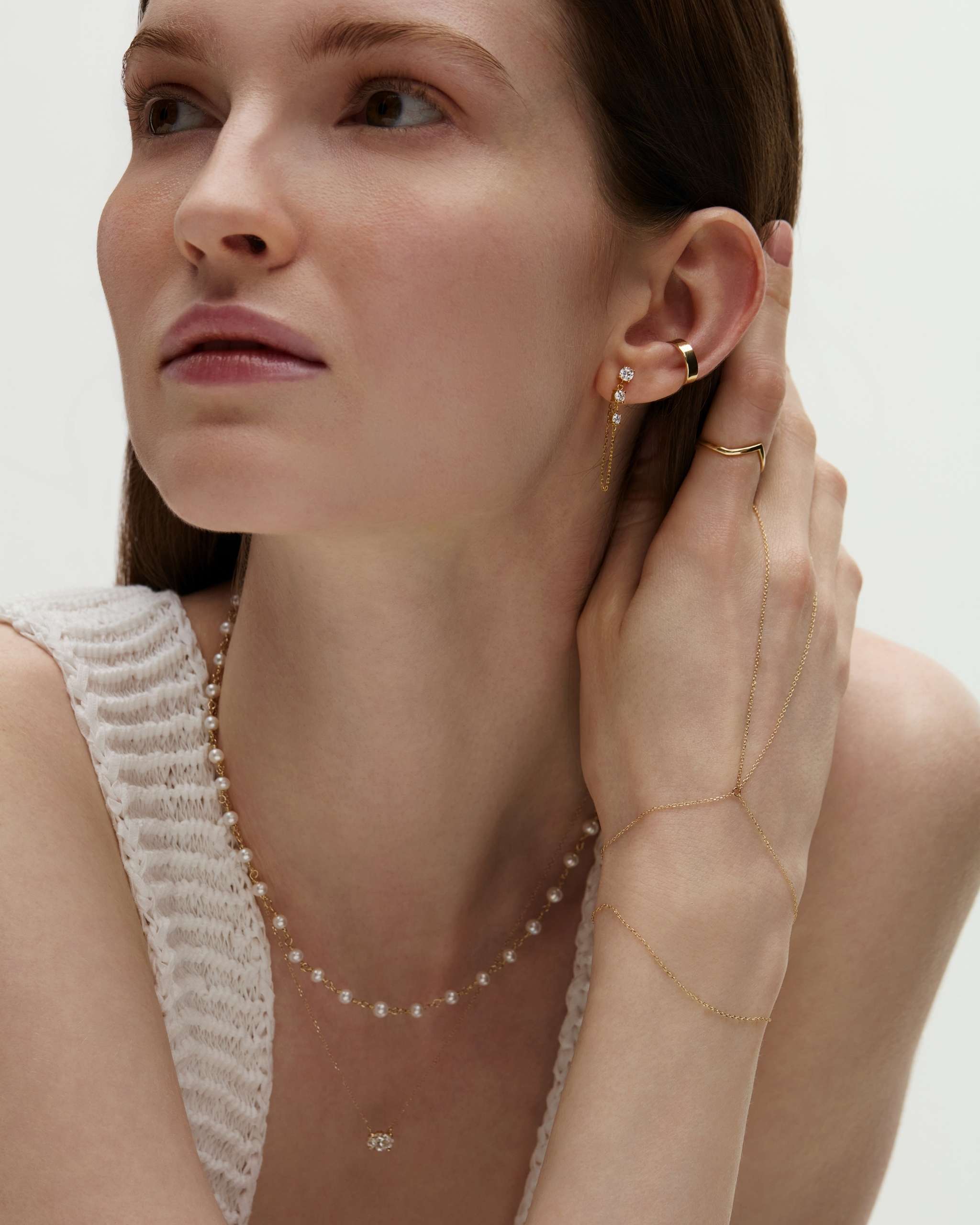 woman resting her hand close to her face wearing a gold ear cuff and diamond chain stud, a dainty gold ring chain bracelet, a 14k gold pearl wrap necklace layered with a longer diamond solitaire necklace on a white background