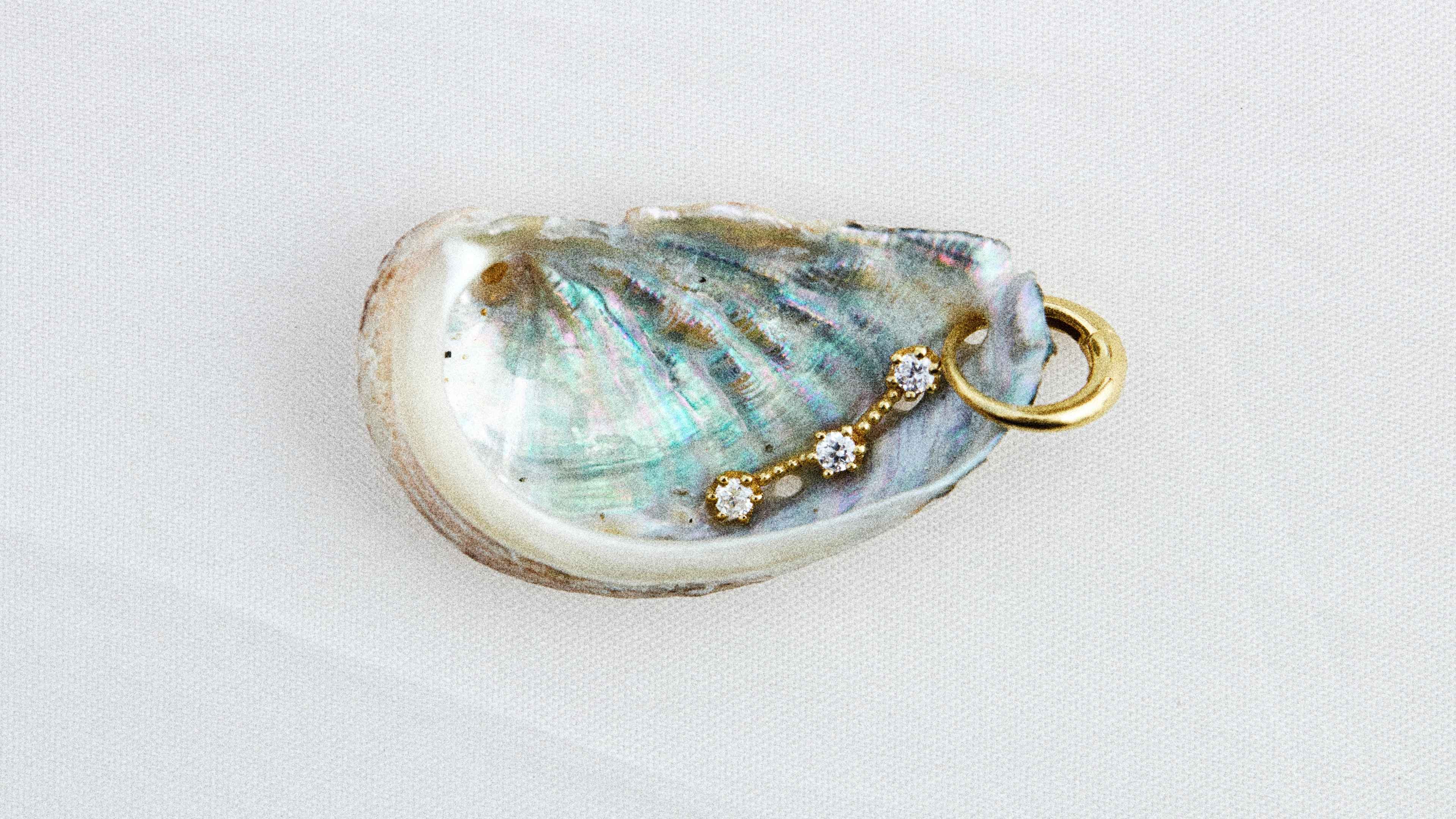 A delicate gold charm with three small diamonds and a gold hoop resting inside an iridescent shell on a light background.