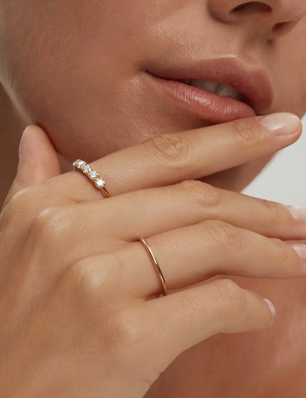 Close-up of a woman resting her hand close to her face wearing a plain yellow gold band styled with another band set with diamonds against a blurred, neutral background. Enea Studio
