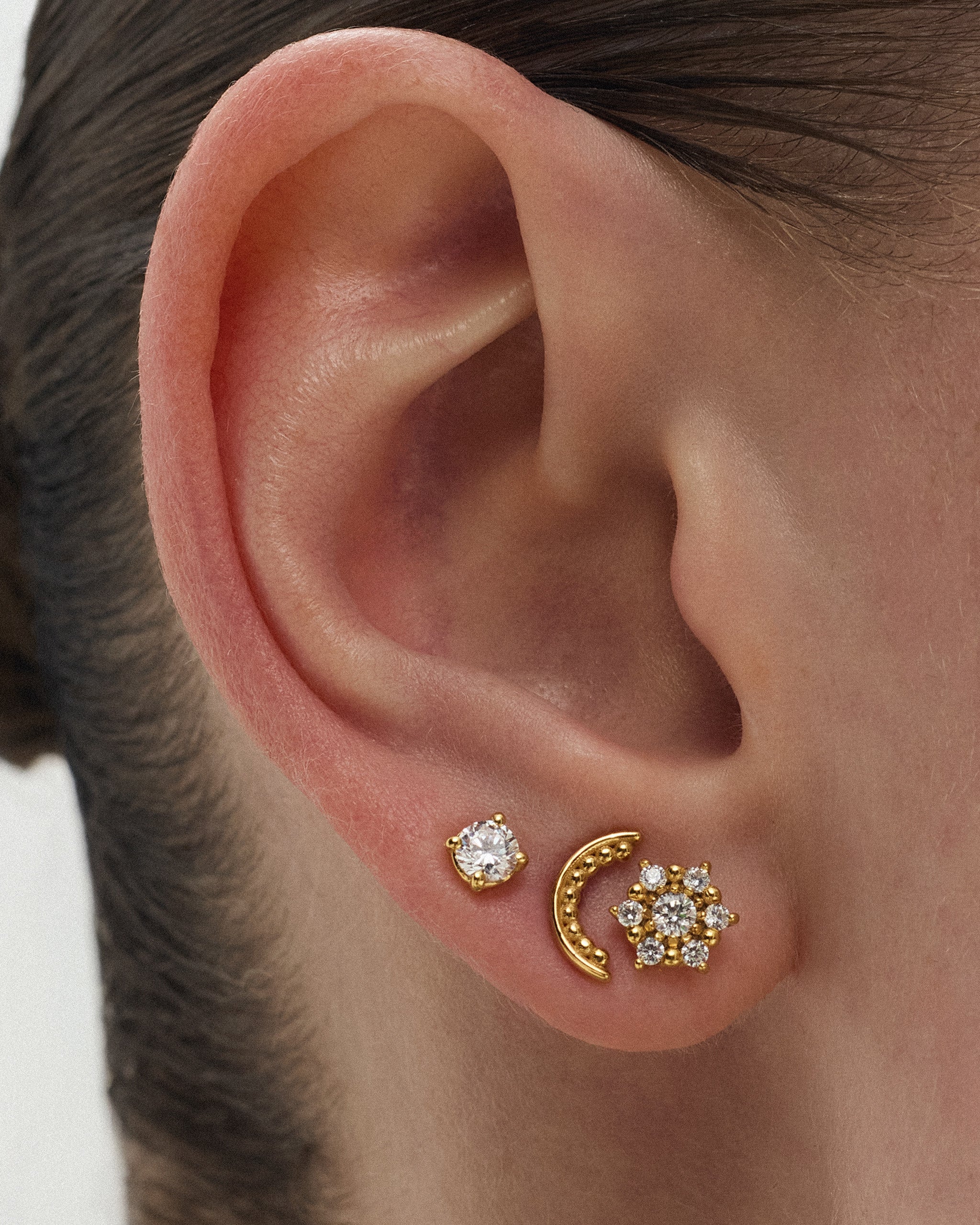 Close up of a woman's ear wearing the Selas Diamond Cluster Stud in 14k yellow gold in the lobe, stacked with a gold crescent stud and a diamond solitaire stud. ENEA Studio