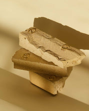 Three stacked rough stones in warm beige and brown tones, each holding a small solid gold ring, photographed against a matching beige background with strong directional shadows.