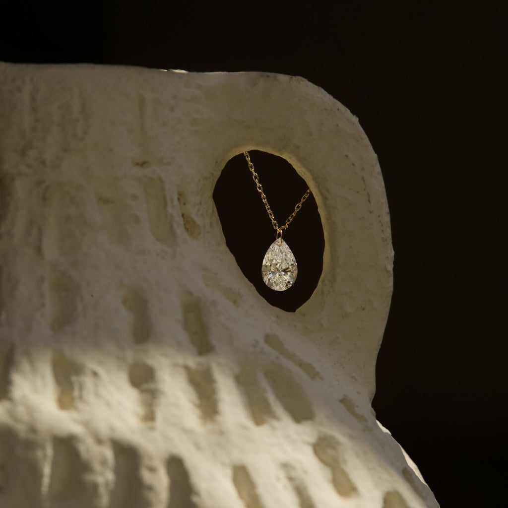 A pear-cut diamond necklace in 14k yellow gold, suspended inside the opening of a textured ceramic sculpture, with soft shadows and black background.