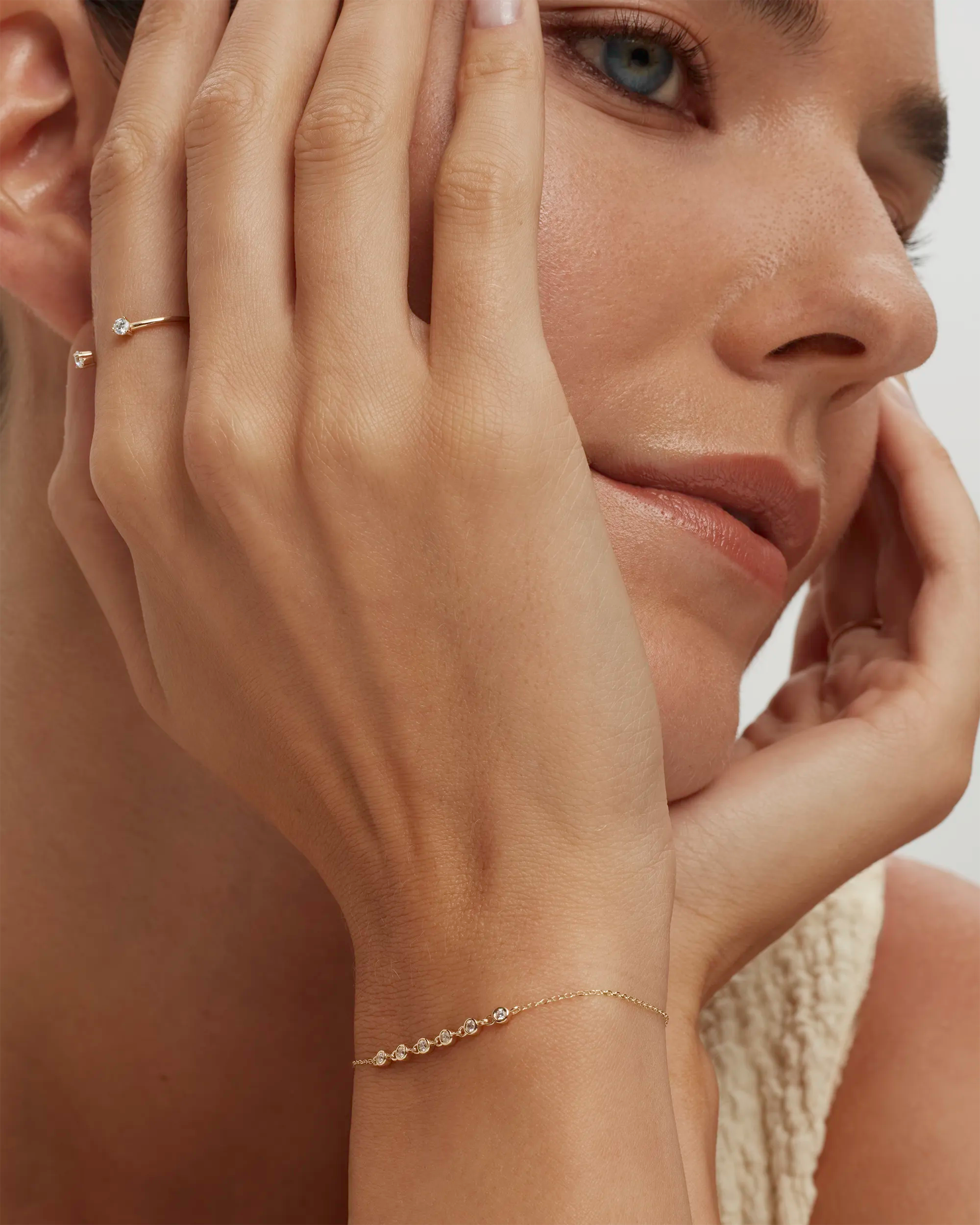 Close-up of a woman's face with one hand on her cheek and a bracelet on her wrist, wearing a ring.