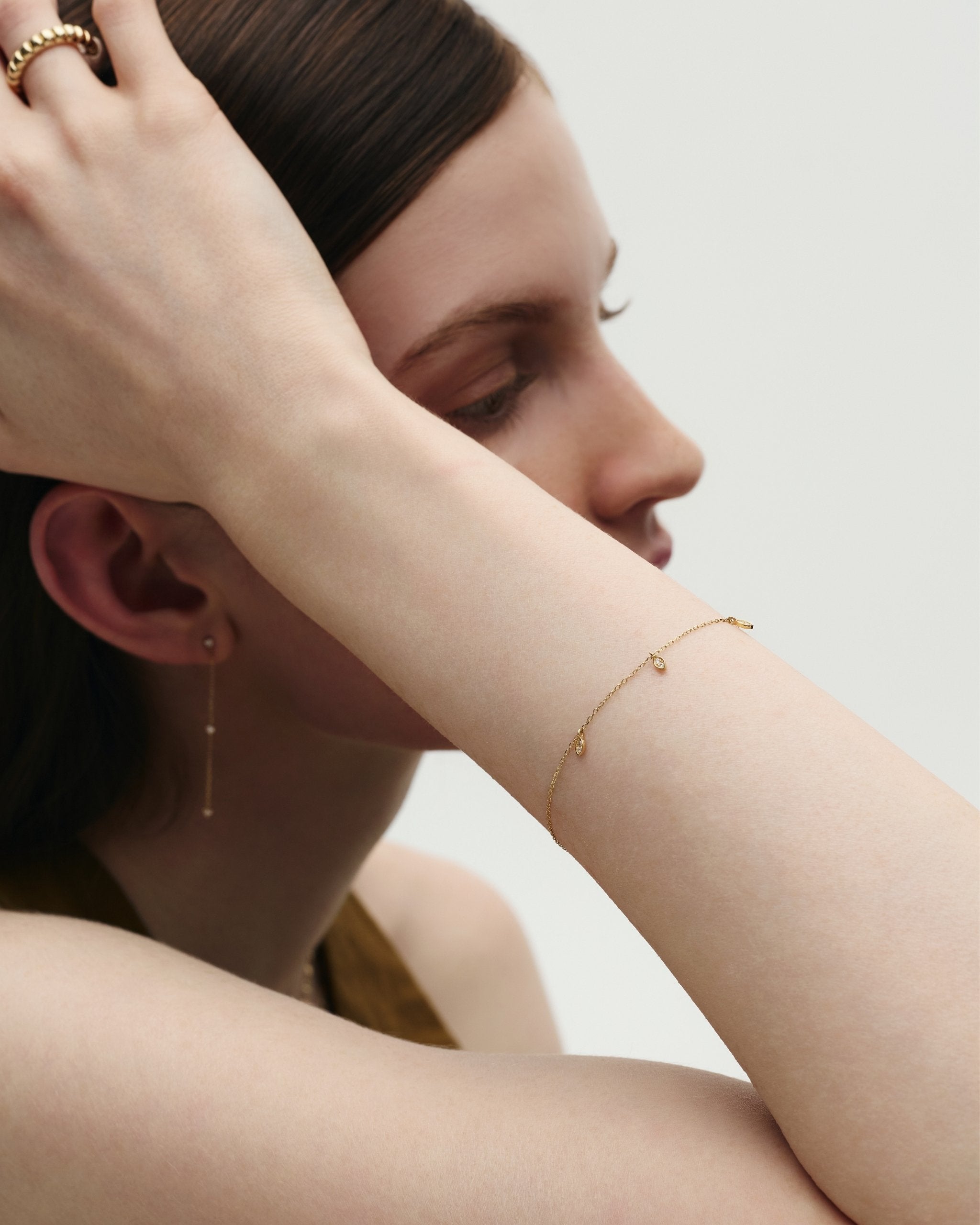 Woman wearing dainty gold jewelry by ENEA including a diamond chain earring, a voluminous gold band, and the Petal Drops Bracelet against a neutral background