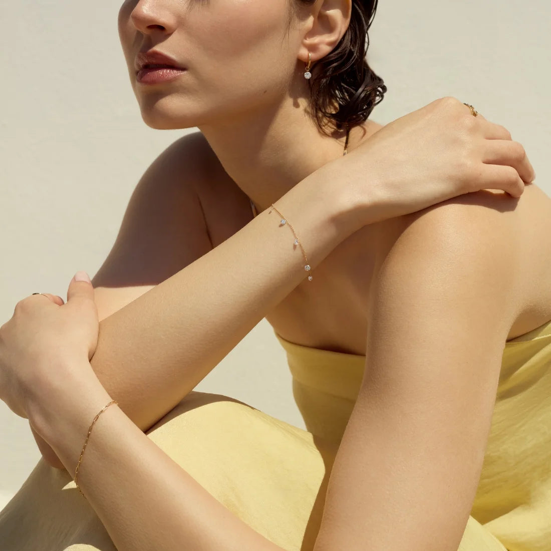 Portrait of a woman wearing a yellow dress styled with 14k gold diamond hoops, a gold paperclip braceelet on one hand, and a diamond station bracelet on the other hand against a neutral background-ENEA