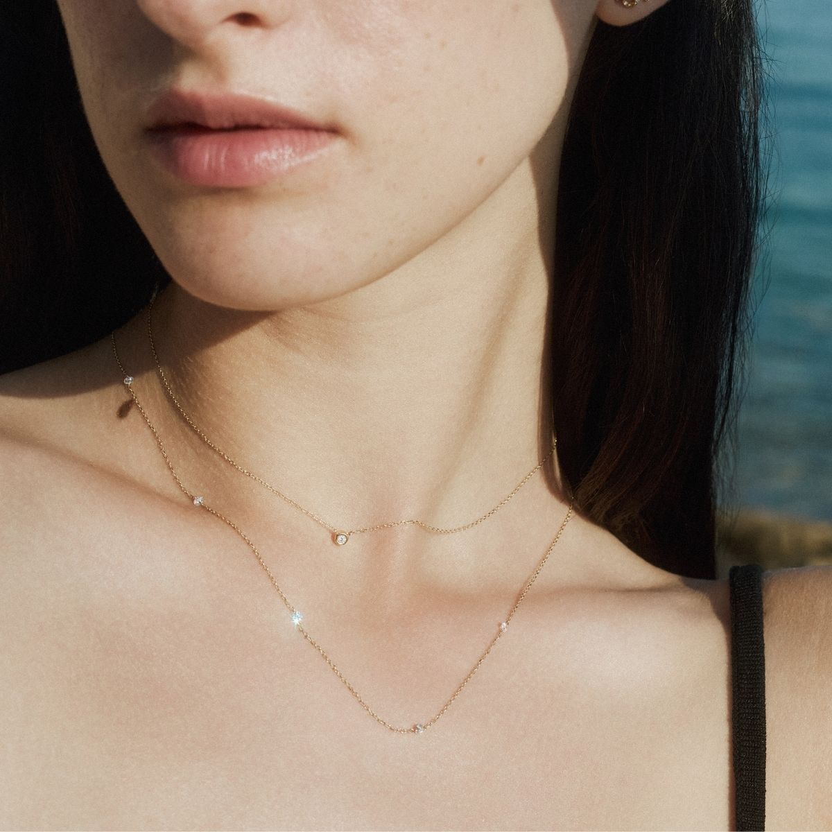 Close-up of a woman wearing a delicate gold diamond solitaire necklace layered with a longer chain accented with small diamonds that glint against blurred background