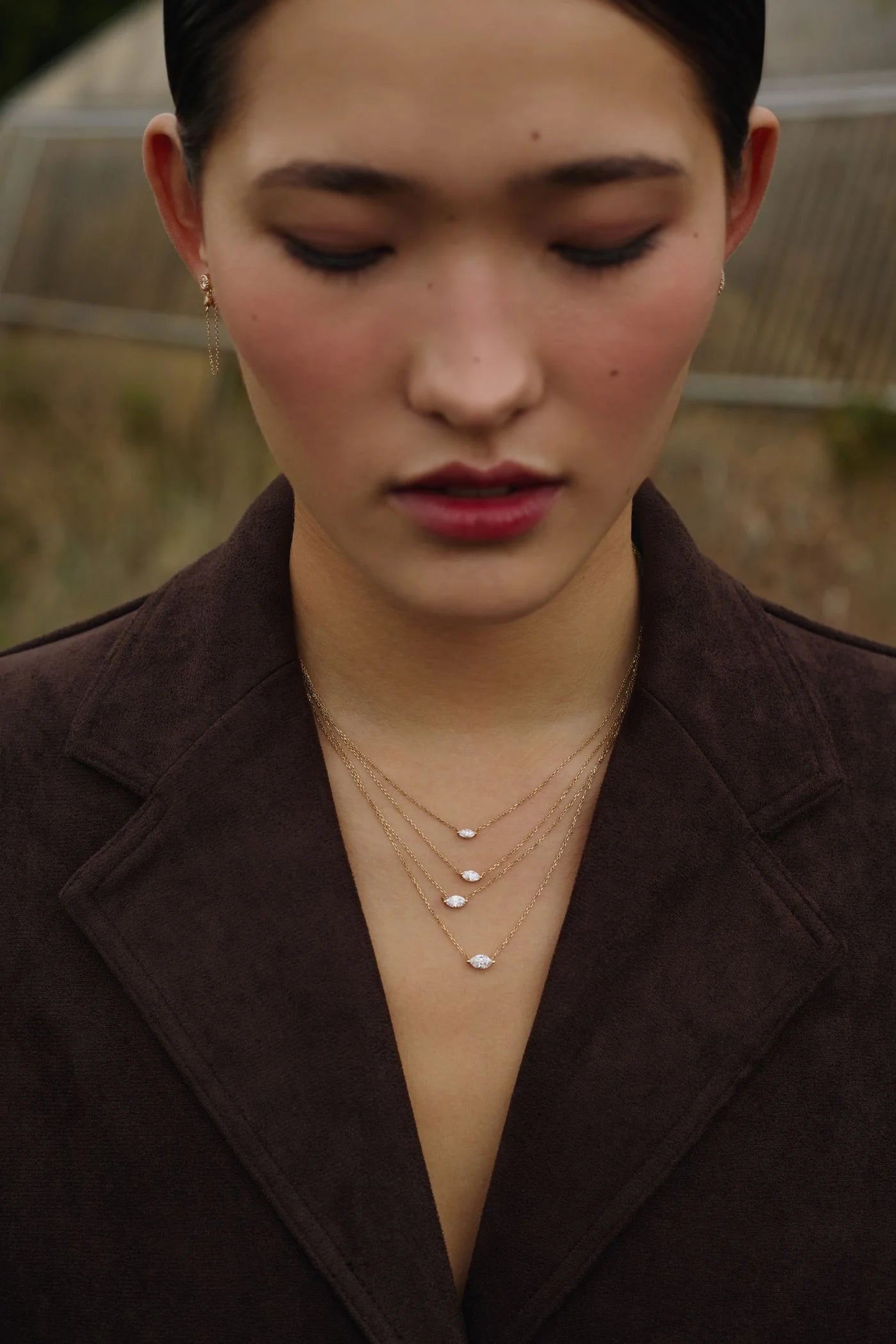 A women wearing a brown velvet coat and four layered diamond solitaire necklaces against a blurred natural background. Enea Studio