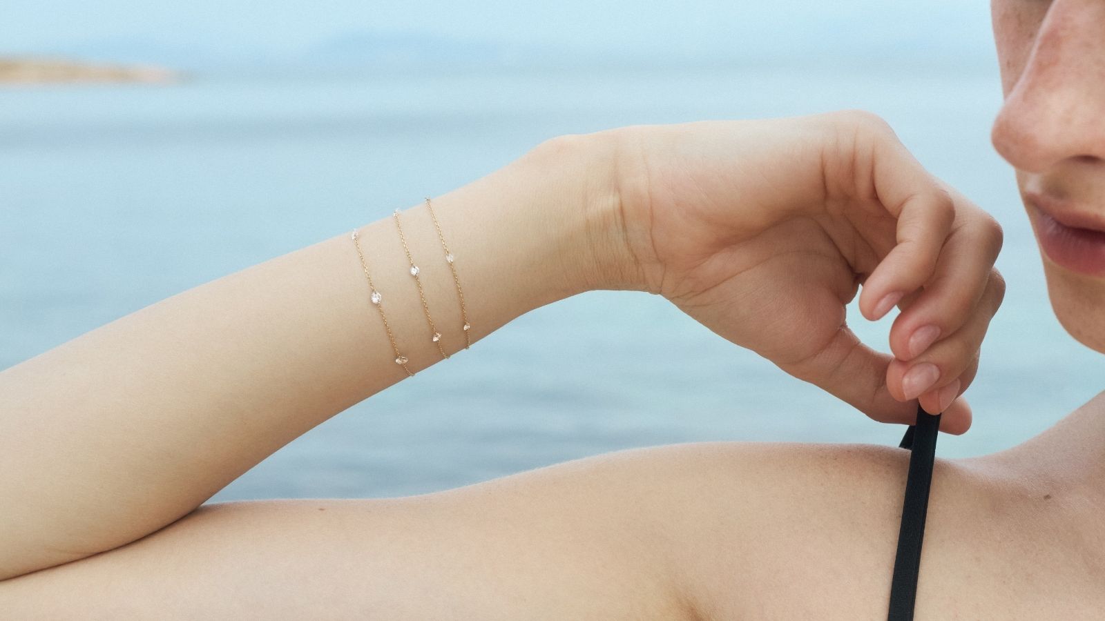 Woman holding the black strap of her bikini above her shoulder with her wrist styled with three dainty diamond chain bracelets by ENEA against a blurred sea background