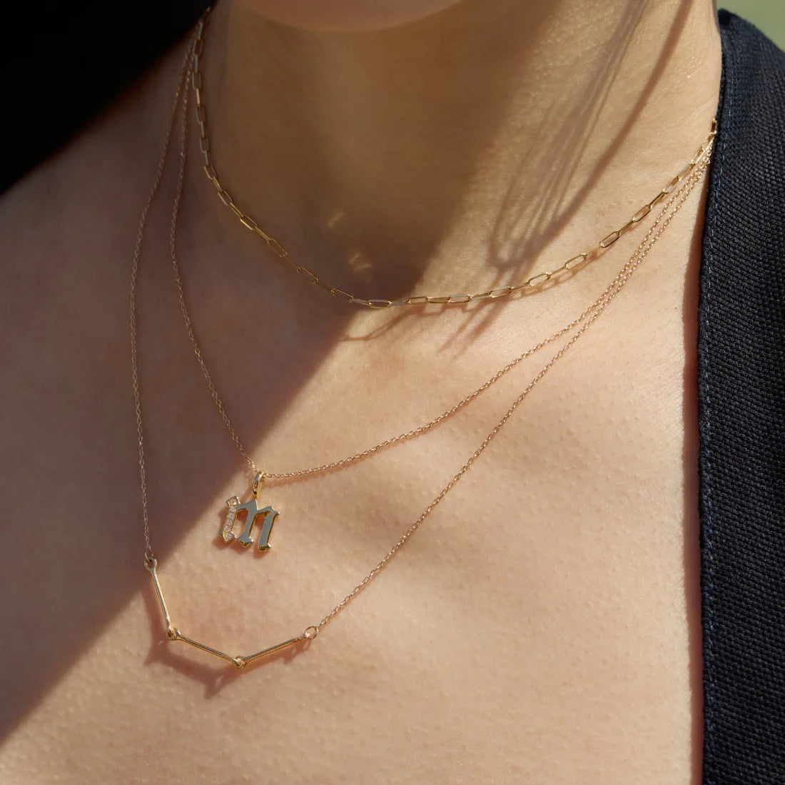 Close-up of a woman wearing a 14k gold chain necklace stacked with a 14k gold initial M pendant encrusted with tiny diamonds, and a long 14k gold necklace with links-ENEA