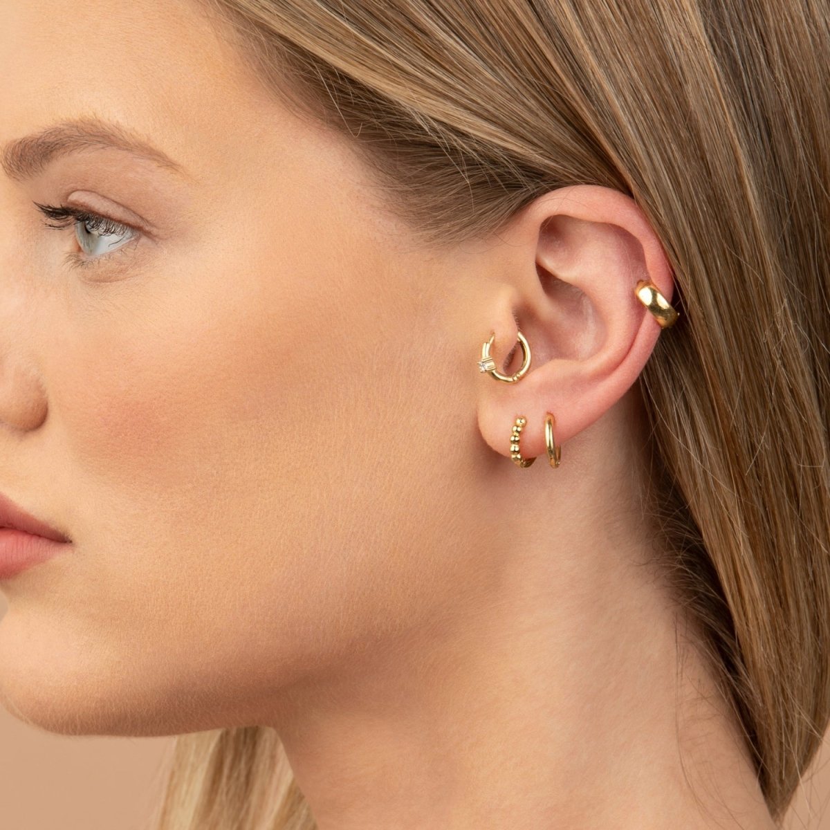 A woman showcasing a 14k yellow gold diamond huggie hoop earring styled with multiple ear piercings, emphasizing a modern and layered look.
