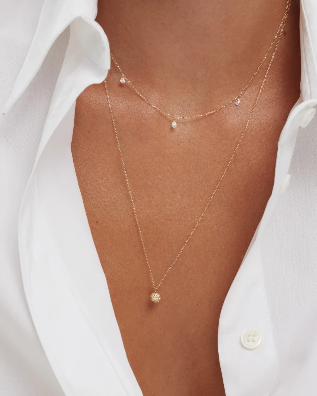 A close-up photo showing a woman's neckline adorned with 2 delicate gold necklaces. One worn close to her collarbone featuring delicate diamonds, and a longer one that features a small sphere pendant accented with small diamonds. She wears a crisp white shirt open at the collar.