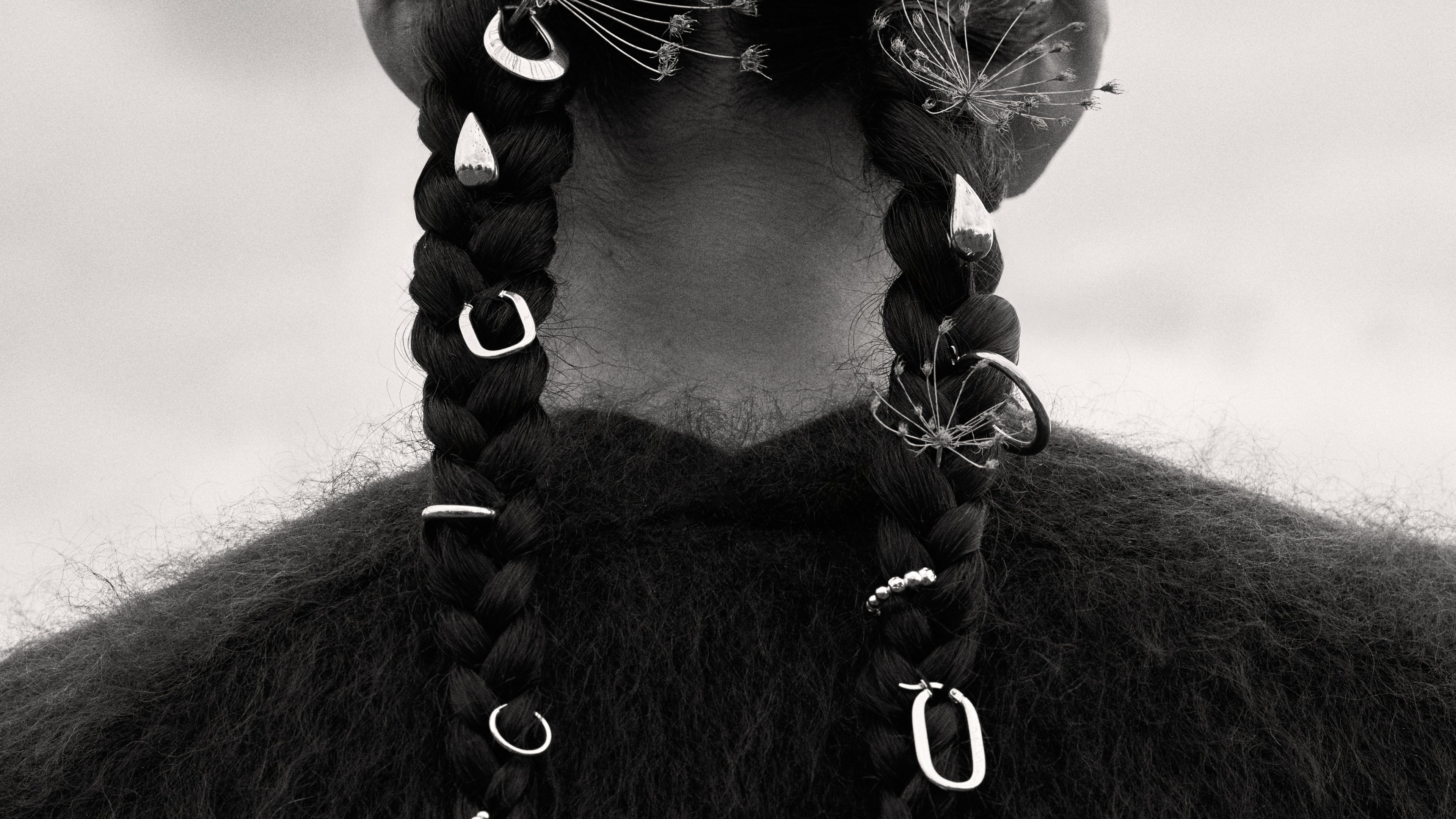 Black and white photo of a woman's back in braided hair adorned with flowers and 14k gold earrings across the 2 braids. — Enea.