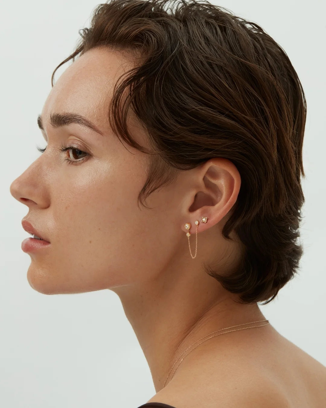 Side photo of a woman wearing delicate 14k solid gold earrings with a dangling chain detail against a neutral background. Enea Studio
