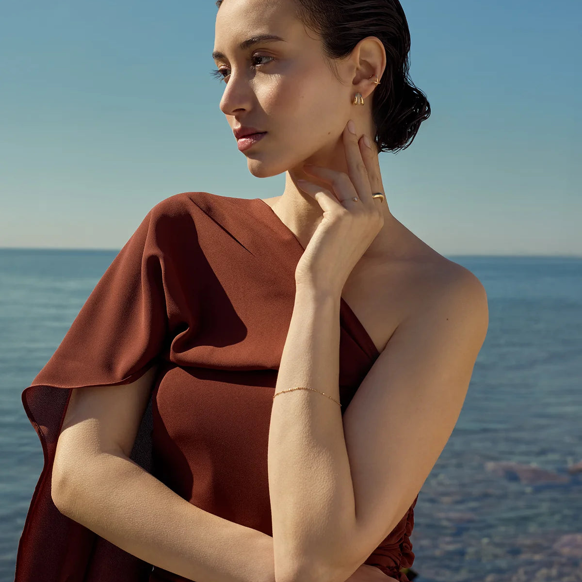 Woman in a rust-colored dress standing by the sea and against clear blue sky wearing delicate gold jewelry from Enea.