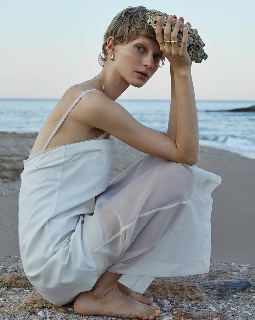 A woman crouches on a sandy beach with the ocean in the background, wearing a flowing white wrap dress and holding a large shell to her forehead.