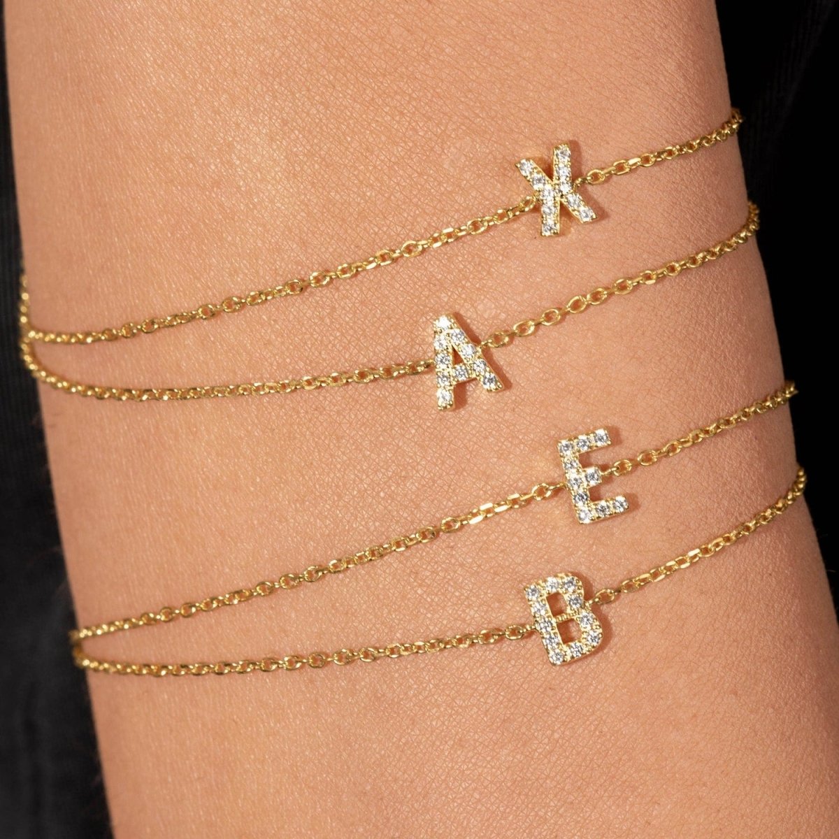 Model’s wrist stacked with solid gold initial bracelets featuring pavé letters A, X, E, and B.
