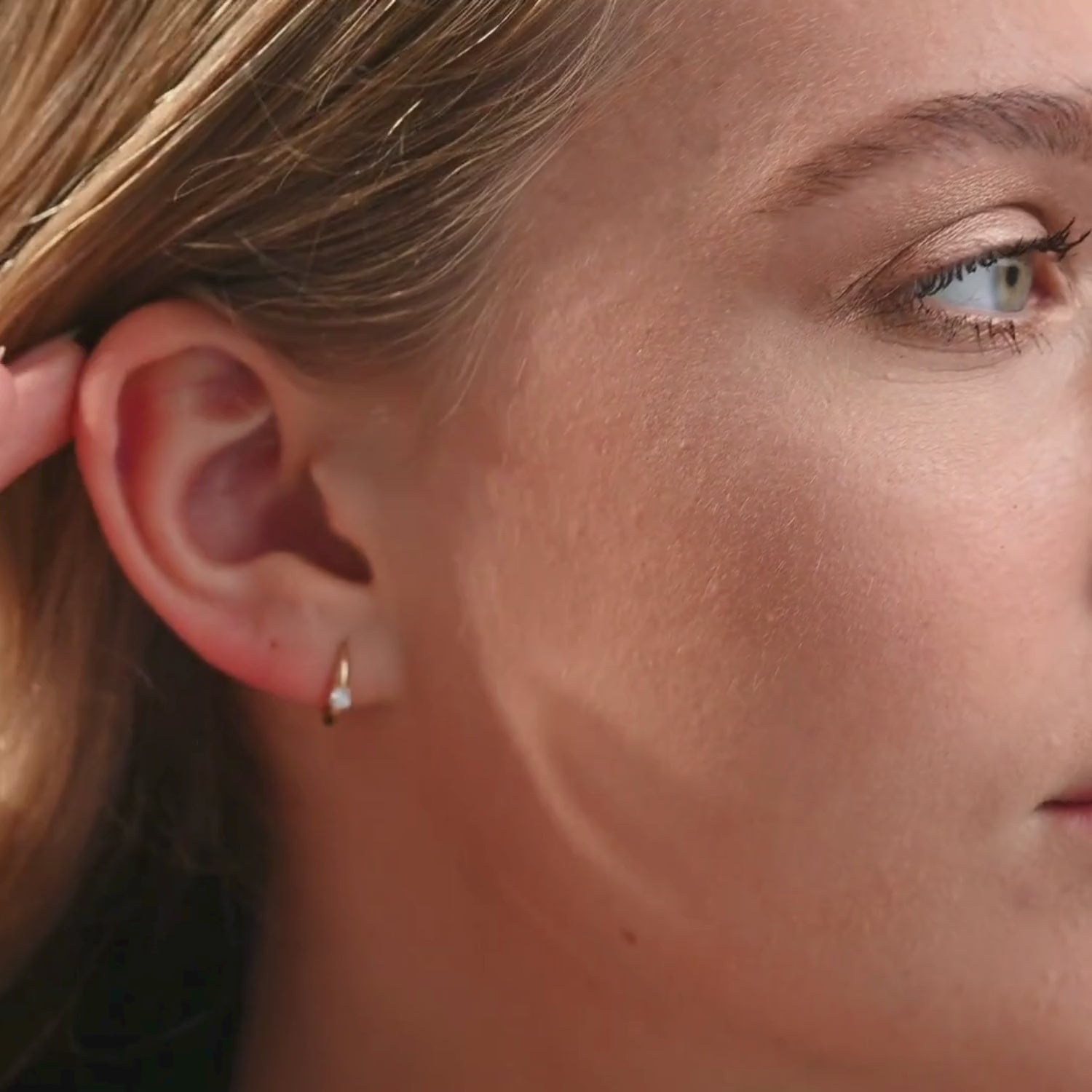 A video of a woman wearing 14k yellow gold diamond huggie hoop earrings, moving naturally to showcase the snug fit and subtle sparkle of the round brilliant-cut diamond.
