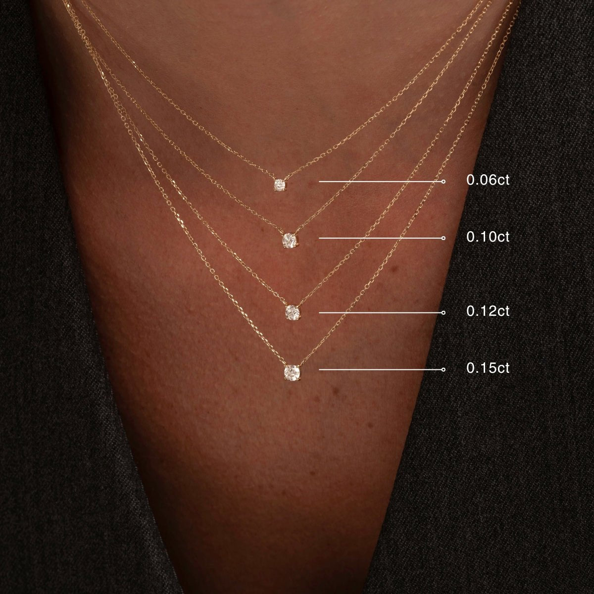 Close-up view of a woman’s chest wearing four 14k gold solitaire diamond necklaces layered vertically, labeled by carat size (0.06ct, 0.10ct, 0.12ct, 0.15ct), against a dark outfit.